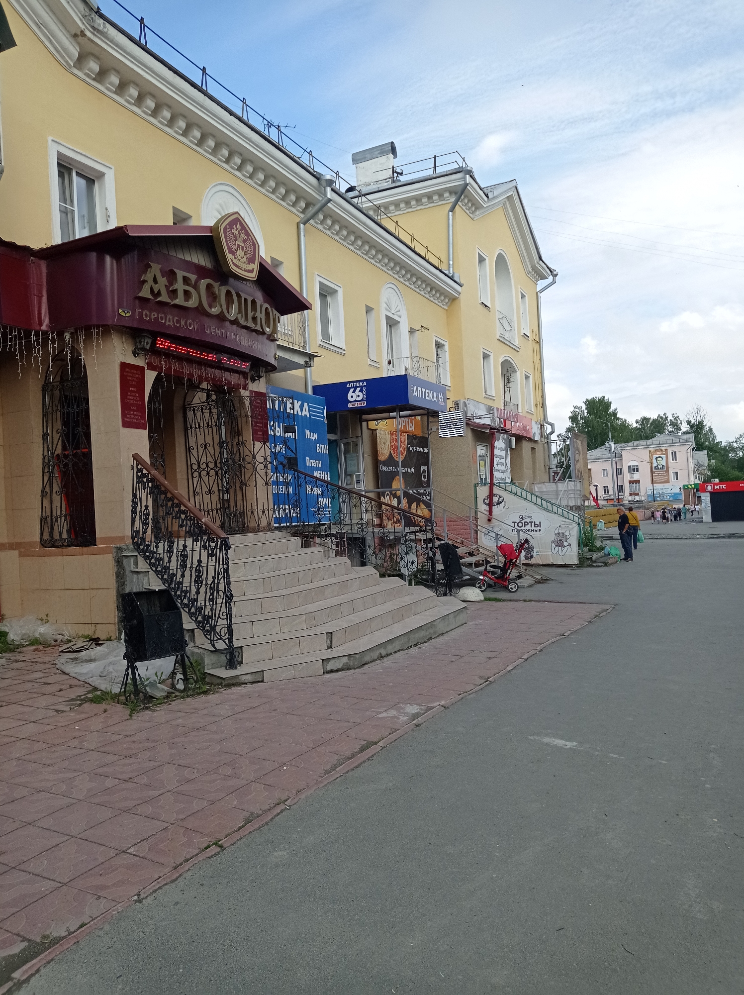 The image captures a street scene in Degtyarsk, Russia, focusing on the facade of a building housing several businesses. The building is painted a light yellow with white architectural detailing along the roofline and around the windows. A prominent burgundy awning with the gold lettering "АБСОЛЮТ" (ABSOLUTE) and "ГОРОДСКОЙ ЦЕНТР НЕДВИЖИМОСТИ" (CITY REAL ESTATE CENTER) is visible on the left. Below this awning, an entrance is protected by ornate black metal gates. To the right of this entrance, a set of wide, light-colored tiled stairs with a black metal railing leads up to another entrance.
Further to the right, a blue sign with white lettering reads "АПТЕКА 66" (PHARMACY 66), indicating a pharmacy. Next to it, a sign advertises "ТОРТЫ" (CAKES) with images of cakes and pastries. More businesses are visible further down the street, with people walking in the background. The street in the foreground is paved with red and gray tiles and asphalt. A red stroller is parked near the stairs, and a trash bin is located at the bottom of the stairs. The sky above is partly cloudy.