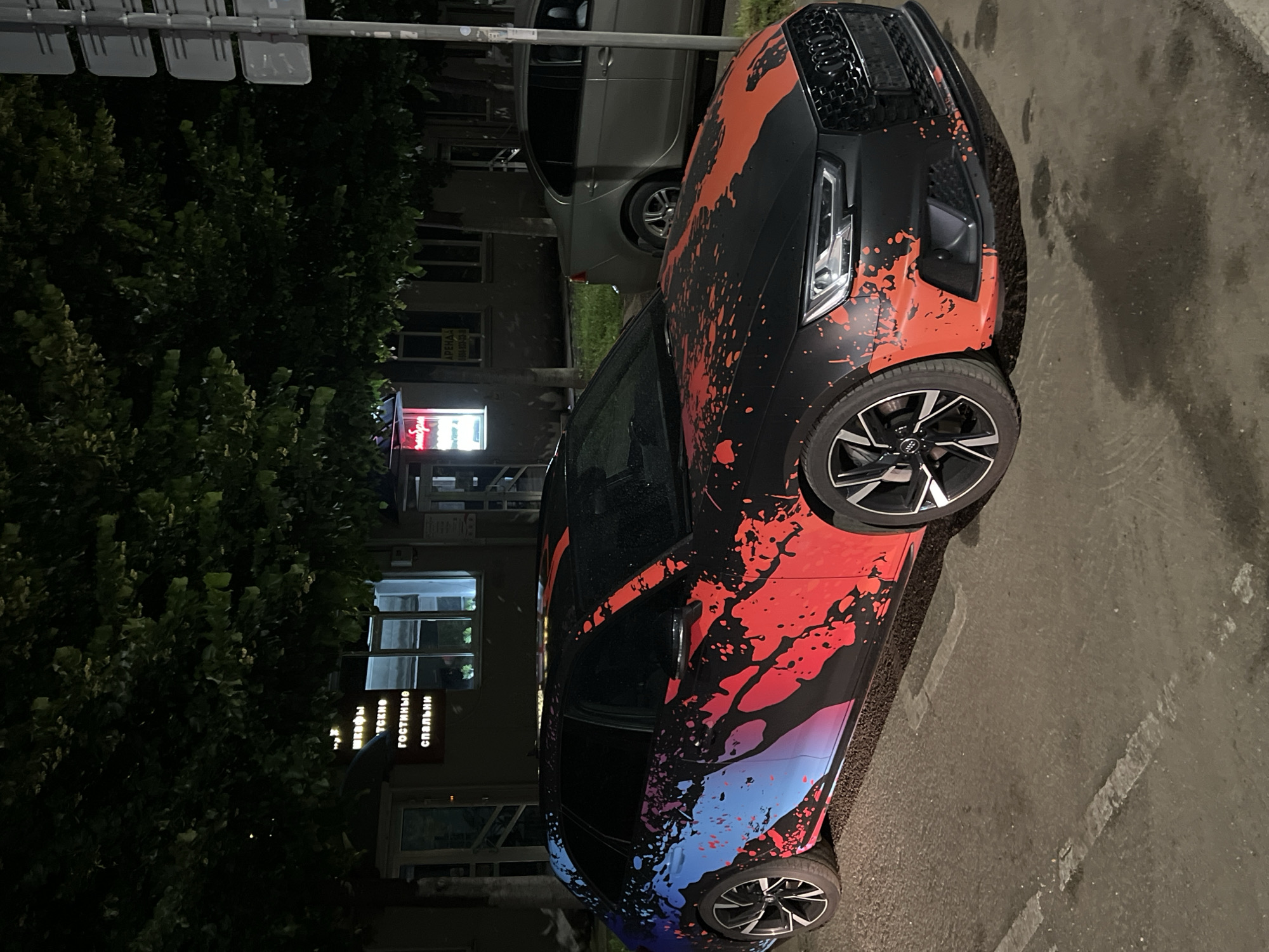 A black Audi A4 Avant is parked on a paved street at night in the city of Krasnodar, Russia. The car is heavily customized with a vibrant, abstract design featuring splashes of red, blue, and pink paint on its black exterior. The car is the primary subject, and it occupies the majority of the frame.
In the background, a light-colored sedan is parked behind the Audi. To the left of the Audi, there is a building with illuminated windows and a sign with Cyrillic text. The building appears to be a commercial establishment. Trees with dense foliage are visible above the car and the building, casting shadows. A street sign is present on the right side of the image. The ground is wet, suggesting recent rain or a spill. No people are visible in the image. The overall atmosphere is urban and nocturnal.