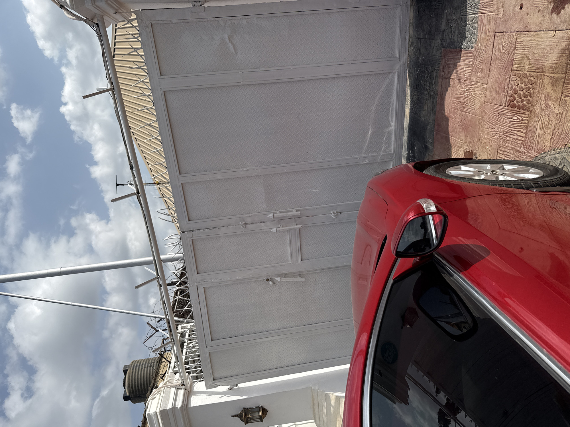 The image shows a red car parked in front of a white metal gate with barbed wire on top. The gate is closed. Behind the gate, a building with a beige roof is visible, along with a water tank. The sky is partly cloudy. The scene appears to be an exterior shot of a property in a residential or commercial area, likely in Abuja, Nigeria, given the presence of a water tank, a common sight in areas with intermittent water supply. The ground is paved with patterned tiles that resemble wood. No people are present in the image. The overall impression is one of security and privacy.