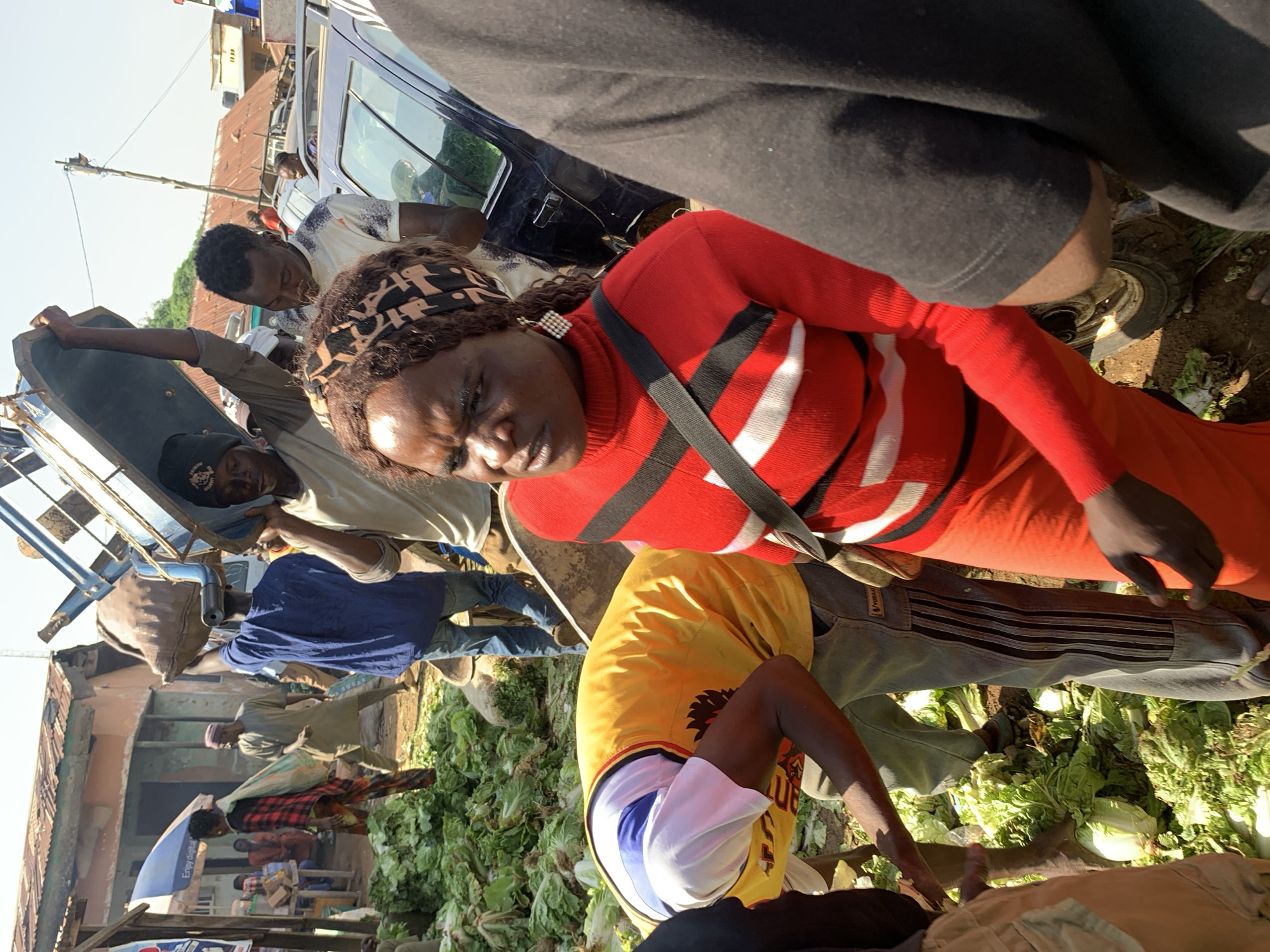 This is a medium shot of a busy outdoor market in Jos, Nigeria, likely a produce market given the large piles of leafy green vegetables on the ground. Several people are present, engaged in various activities related to the market. In the foreground, a woman wearing a red turtleneck sweater with white stripes and an orange skirt is looking to her left with a concerned expression. She has a dark headscarf and earrings. To her left, a person wearing a yellow vest over a white shirt is bending over, appearing to sort or handle the vegetables. Behind them, a man in a blue shirt and jeans is walking away, and another man in a light-colored shirt is carrying a large metal cart or bin on his head. In the background, more people, market stalls, and vehicles are visible, suggesting a bustling commercial environment. The ground is dirt and littered with vegetables. The overall impression is one of active commerce in an informal urban setting.