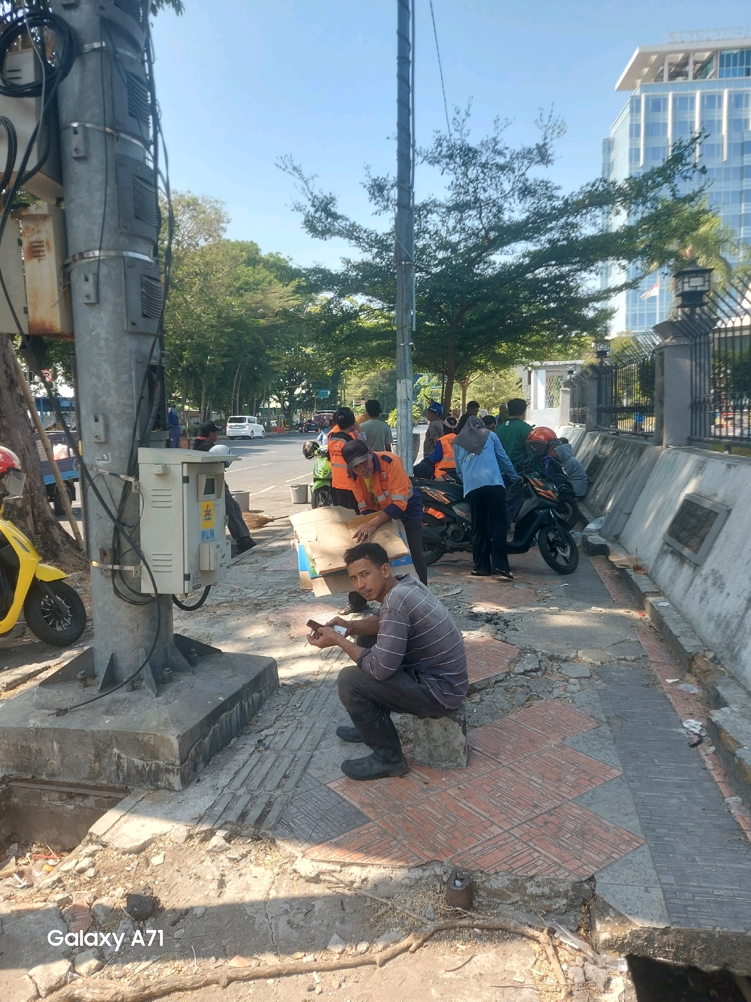 The image depicts a street scene in Makassar, Indonesia, possibly in a commercial or industrial area, indicated by the modern glass building in the background. In the foreground, a man in a striped shirt and dark pants is sitting on a concrete block on a damaged sidewalk. He is holding a small object, possibly a phone or a tool, in his hands. Several other individuals, some wearing orange work vests, are gathered around motorcycles and what appears to be utility equipment. A large metal pole with electrical boxes is prominently positioned to the left of the frame. The sidewalk is in disrepair, with broken concrete and exposed earth. A yellow scooter is parked to the far left. The overall environment suggests a public space undergoing some form of work or maintenance.