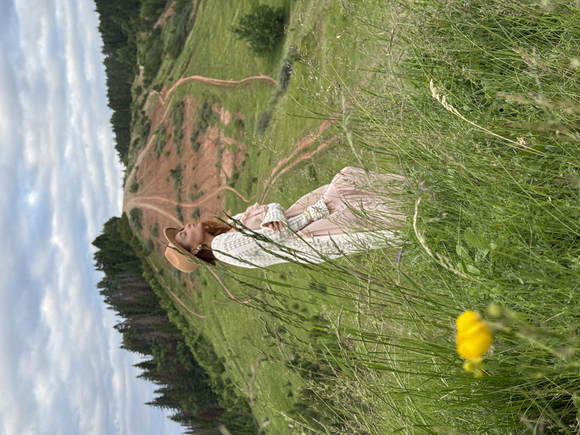 A woman wearing a wide-brimmed hat, a white open-knit cardigan, and a light pink dress stands in a field of tall grass. She is looking upwards with her eyes closed, her head tilted back. The background features a grassy hillside with a winding dirt path, dotted with trees and a patch of exposed red earth. The sky above is overcast with scattered clouds. The location is Olkhovka, Russia.
