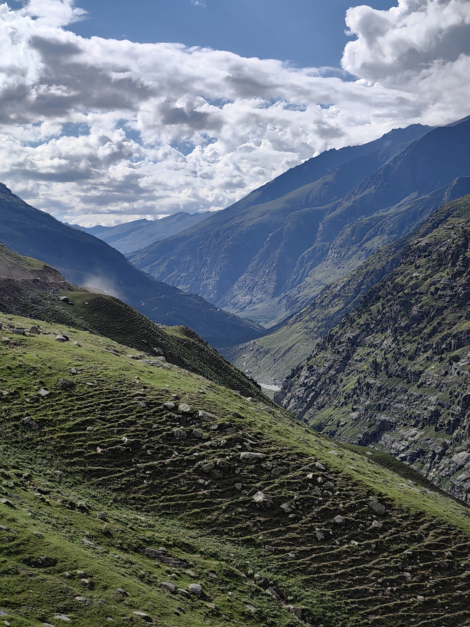 The image captures a panoramic view of a mountainous valley in the vicinity of Bashisht, India. The foreground features a steep, grassy slope with visible terracing, suggesting agricultural use. Scattered rocks dot the green landscape. A dirt road curves along the upper edge of this slope, and a faint plume of dust rises from it, indicating recent activity.
Beyond the foreground, the valley widens, revealing more rugged, rocky mountainsides covered in patches of green vegetation. The mountains recede into the distance, their peaks forming layered silhouettes against a partly cloudy blue sky. The clouds are dramatic, with bright white cumulus formations contrasting with darker, shadowed areas. A narrow river or stream is visible in the depths of the valley, winding its way through the mountainous terrain. The overall impression is one of a remote, natural landscape with signs of human presence through the road and terracing.