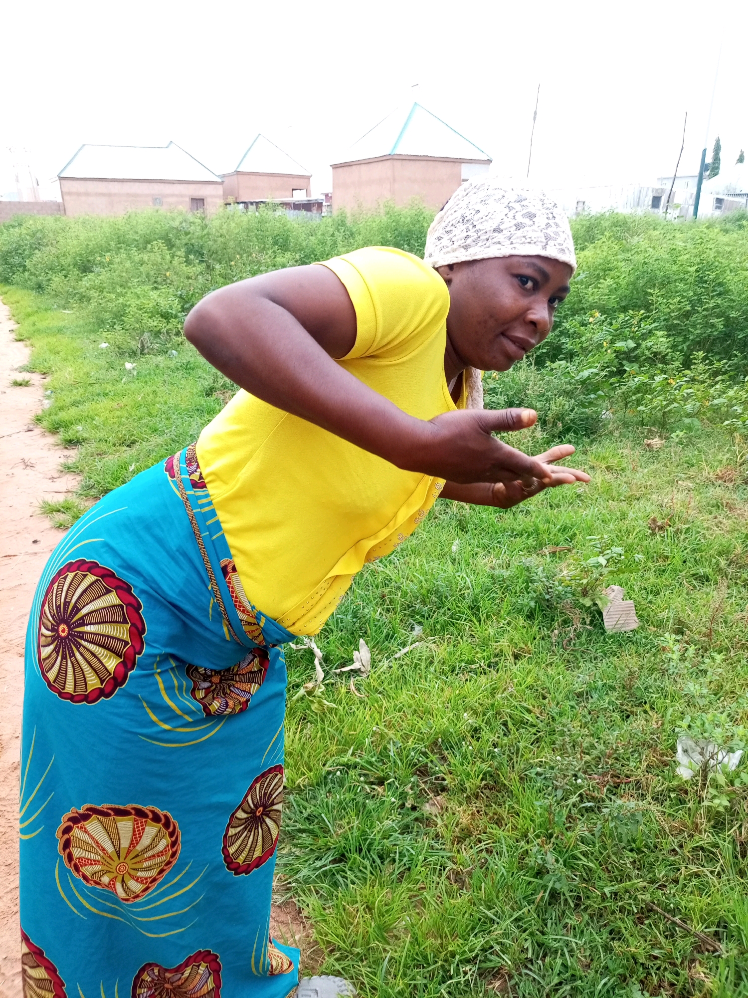 A woman wearing a yellow t-shirt and a patterned blue skirt is bending over in a grassy area. She is looking towards the camera with her hands outstretched. In the background, there are several buildings with light-colored roofs and green foliage. The ground is a dirt path on the left and grass on the right. The scene appears to be in Tempure, Nigeria.