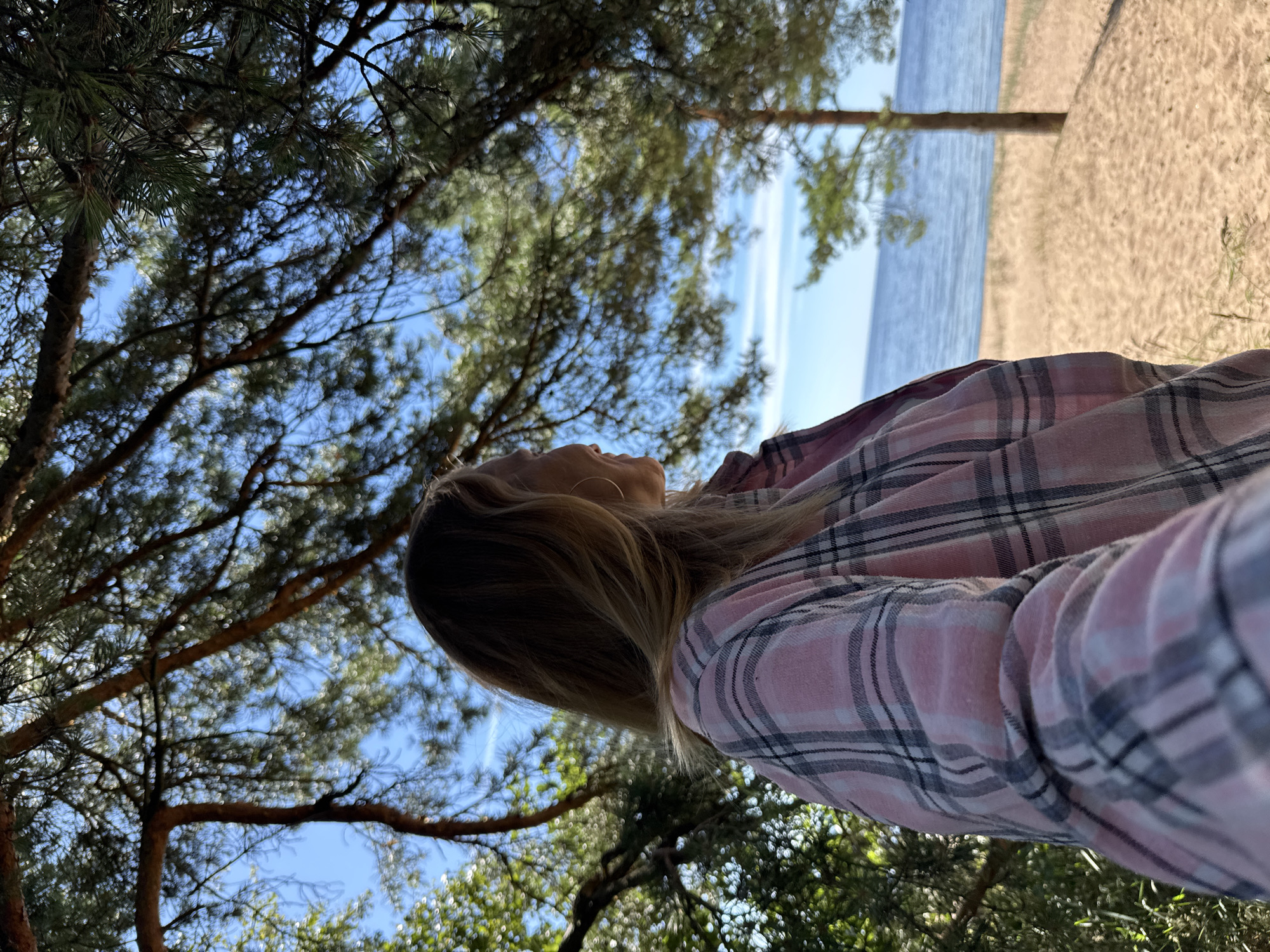 A young woman is positioned in the foreground, facing away from the camera and looking to the right, towards a body of water. She has long, blonde hair and is wearing a pink and gray plaid shirt. She is standing among trees, with pine needles and branches partially obscuring the view of the sky. The background reveals a sandy beach and a calm blue body of water, likely a lake or sea, under a clear blue sky with some wispy clouds. A single pine tree stands on the beach to the right. The overall scene suggests a natural outdoor setting, possibly a park or a coastal area. The location is identified as Zelenaya Roshcha, Russia.