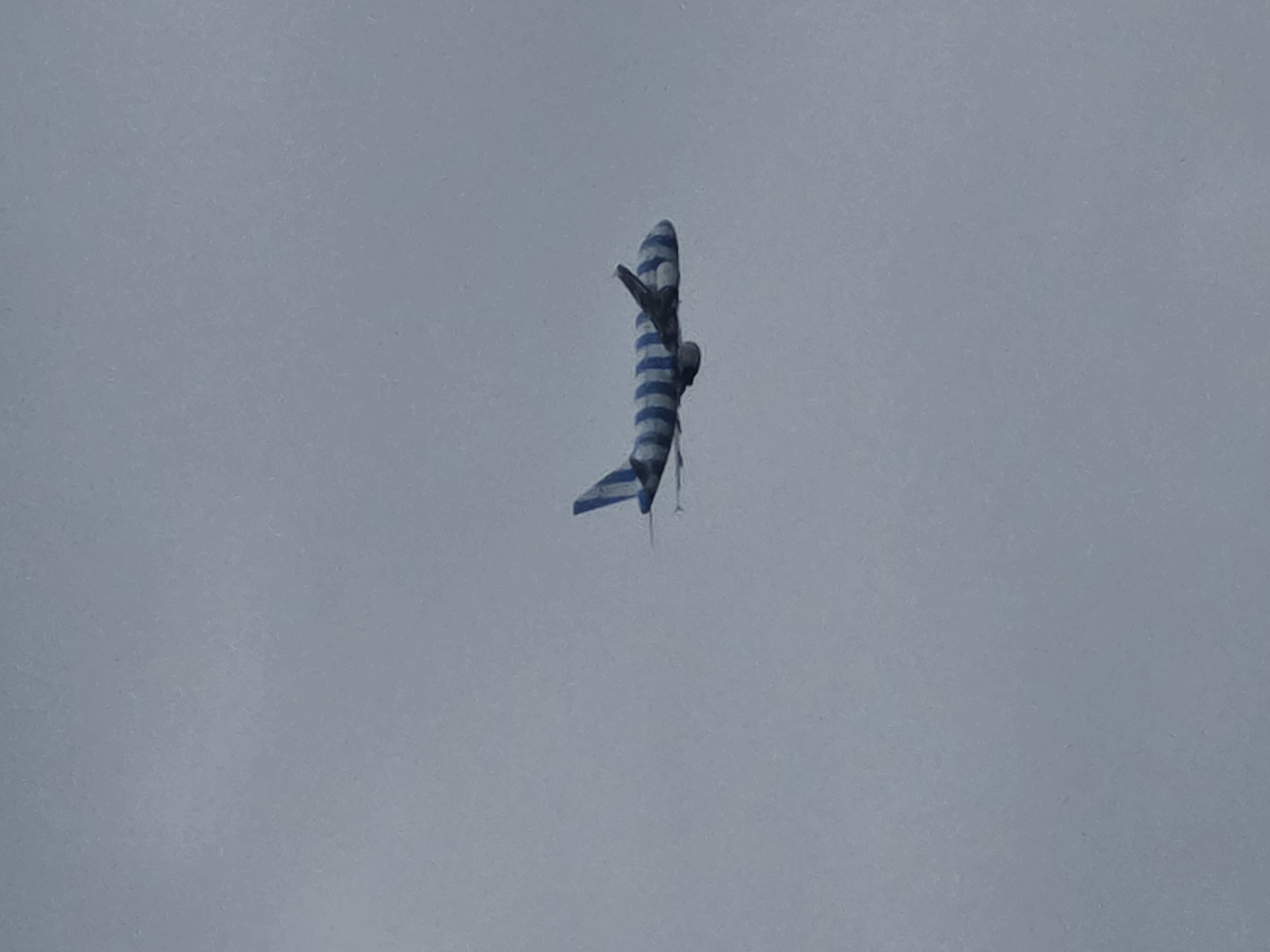 An airplane with a blue and white striped livery is flying in a hazy, overcast sky over Kelsterbach, Germany. The aircraft is visible in profile, with its wings extended and engines mounted beneath them. The sky is a uniform light gray, suggesting a cloudy or foggy day. No other objects or people are discernible in the image. The focus is solely on the airplane against the muted sky.