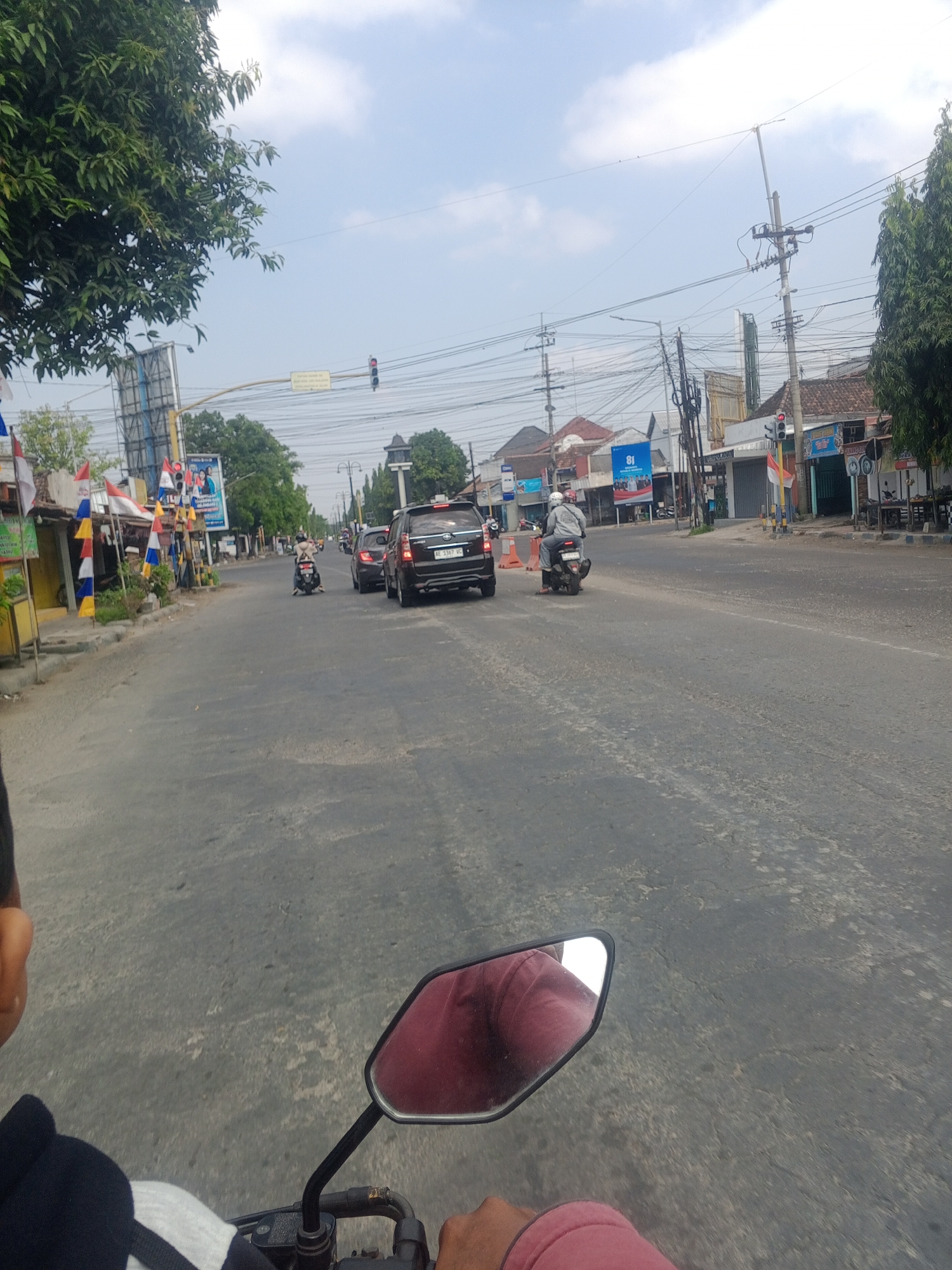 The image depicts a street scene in Kadipaten, Indonesia, on a cloudy day. In the foreground, the perspective is from someone riding a motorcycle, with a red jacket reflected in the side mirror. The road is asphalt, with some wear and tear. A black SUV with license plate AE 3367 WC is traveling ahead. A person on a scooter is also visible in the mid-ground, wearing a white helmet and grey jacket. On the left side of the road, there are several small shops and stalls, decorated with colorful flags. Traffic lights and utility poles with numerous wires are present along the street. In the background, more buildings and trees are visible. The overall scene suggests a typical urban or semi-urban environment in Indonesia with moderate traffic.