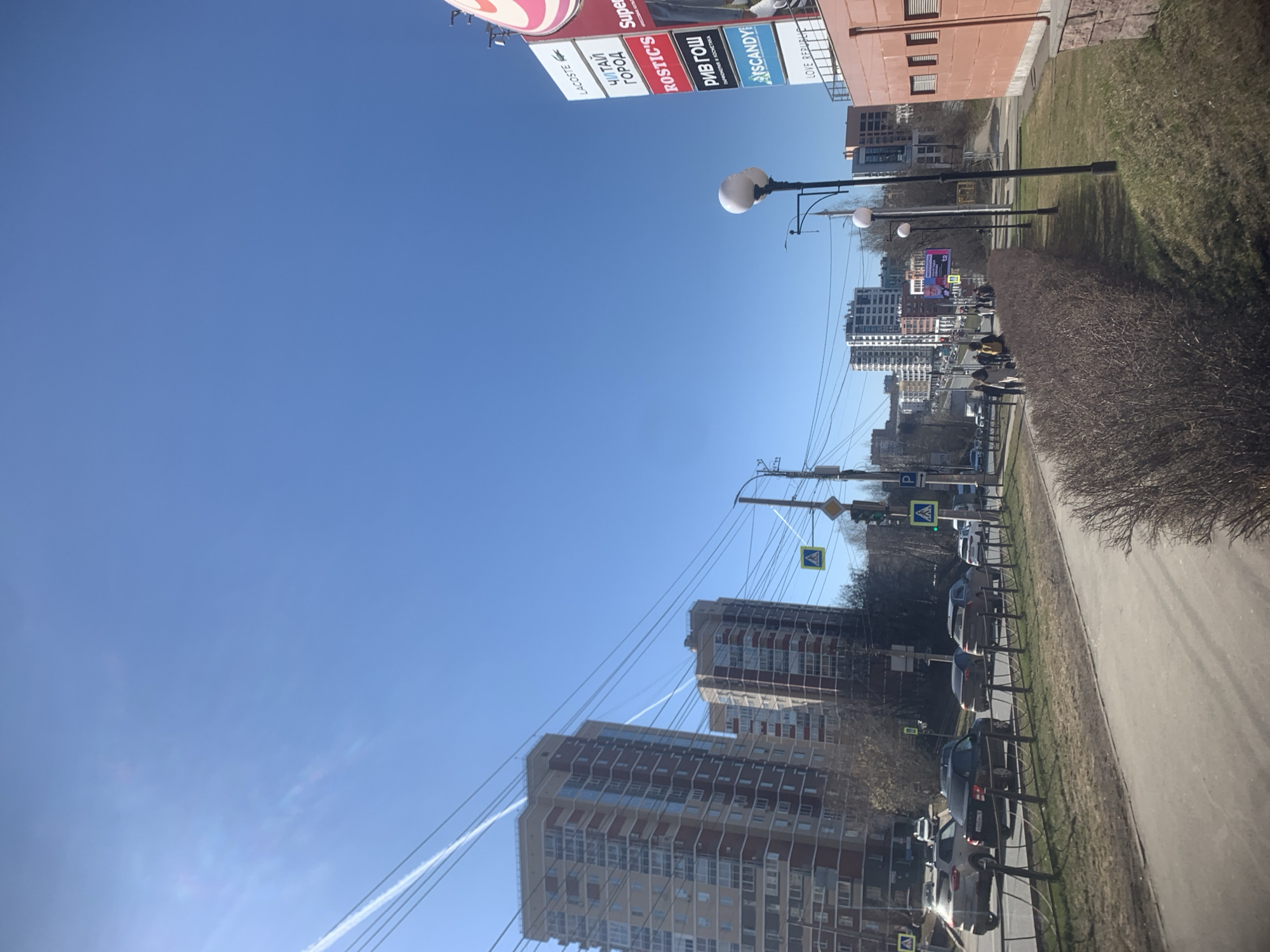 This image captures a bright, sunny urban street scene in Izhevsk, Russia, viewed under a clear blue sky. The time of day appears to be daylight, with the sun casting clear illumination across the environment.
In the foreground, a paved sidewalk runs along the right side, separated from a strip of dormant, brown bushes and grass by a low metal fence. Several cars are parked along a road visible to the left of the sidewalk. In the middle ground, a few pedestrians are scattered, walking on the sidewalk. Numerous streetlights with spherical white lamps are positioned along the street.
The urban landscape in the background consists of several multi-story buildings, including modern residential towers with numerous windows, and an older, reddish-brown commercial or residential structure. Various street signs are present, notably yellow diamond-shaped pedestrian crossing signs and a blue "P" parking sign.
A striking feature in the upper left is a large, vertical advertising sign displaying multiple brand logos. The visible text on this sign, reading from top to bottom, includes "LACOSTE", "ЧИТАЙ ГОРОД" (Chitai Gorod, meaning "Read City"), "ROSTIC'S" (a brand name for KFC in Russia), "РИВ ГОШ" (Rive Gauche), "SCANDY", and "LOVE REPUBLIC".
The blue sky contains a faint white contrail stretching across it. A dense network of overhead wires, likely for power or telecommunications, crisscrosses the frame, adding to the urban complexity of the scene. The overall impression is a typical, bustling city street on a clear day.
