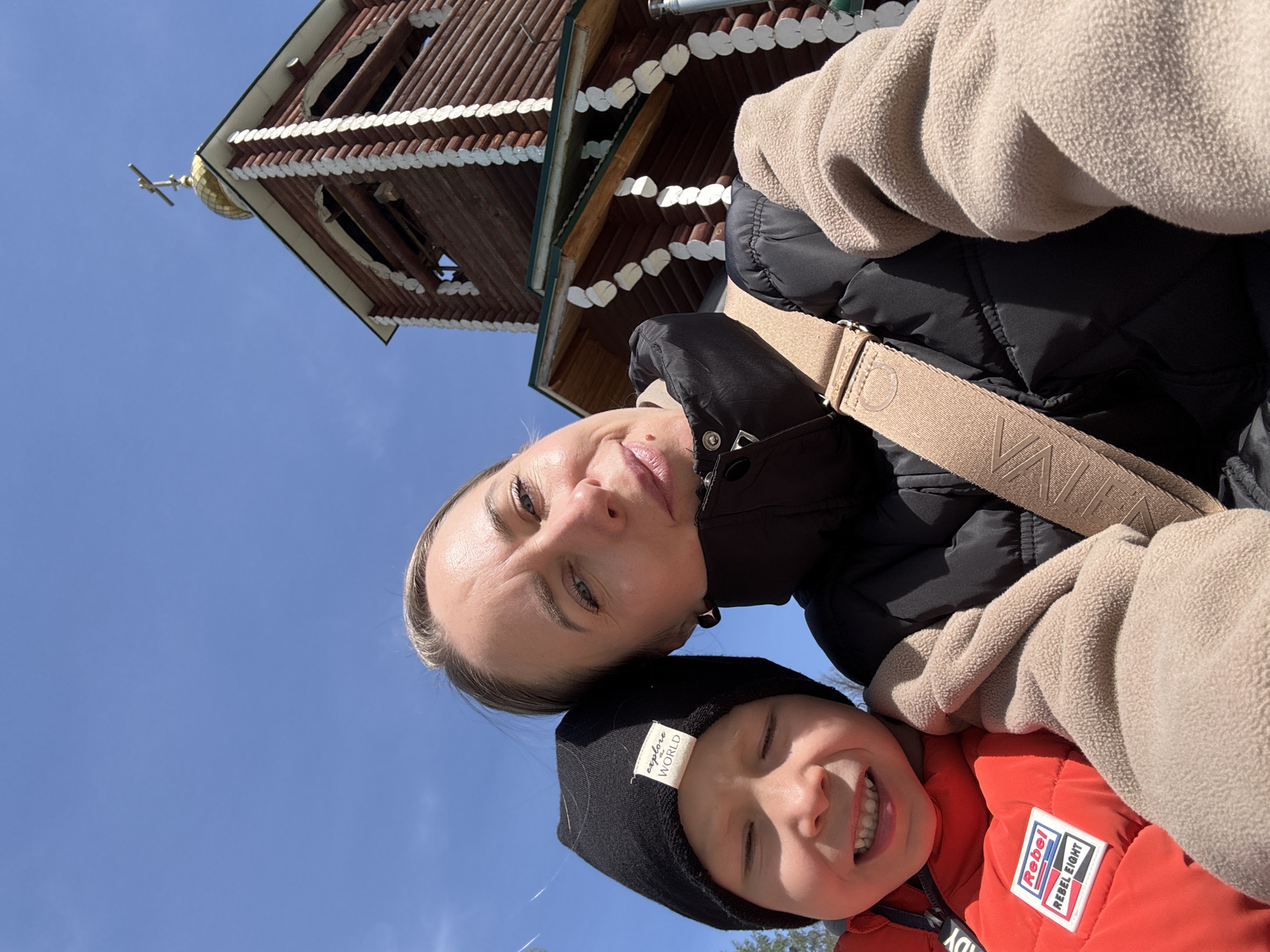 A woman and a young child, possibly a son, are taking a selfie. They are outdoors under a blue sky, with a wooden structure in the background that features white trim and a gold dome with a cross on top. The wooden structure might be the bell tower of the Saint George Church in Chaikovsky, Perm Krai, Russia. The woman is wearing a black jacket, and a beige bag strap is visible across her body. The child is wearing a black hat and an orange puffer jacket with a logo patch visible on the front. Both are smiling.