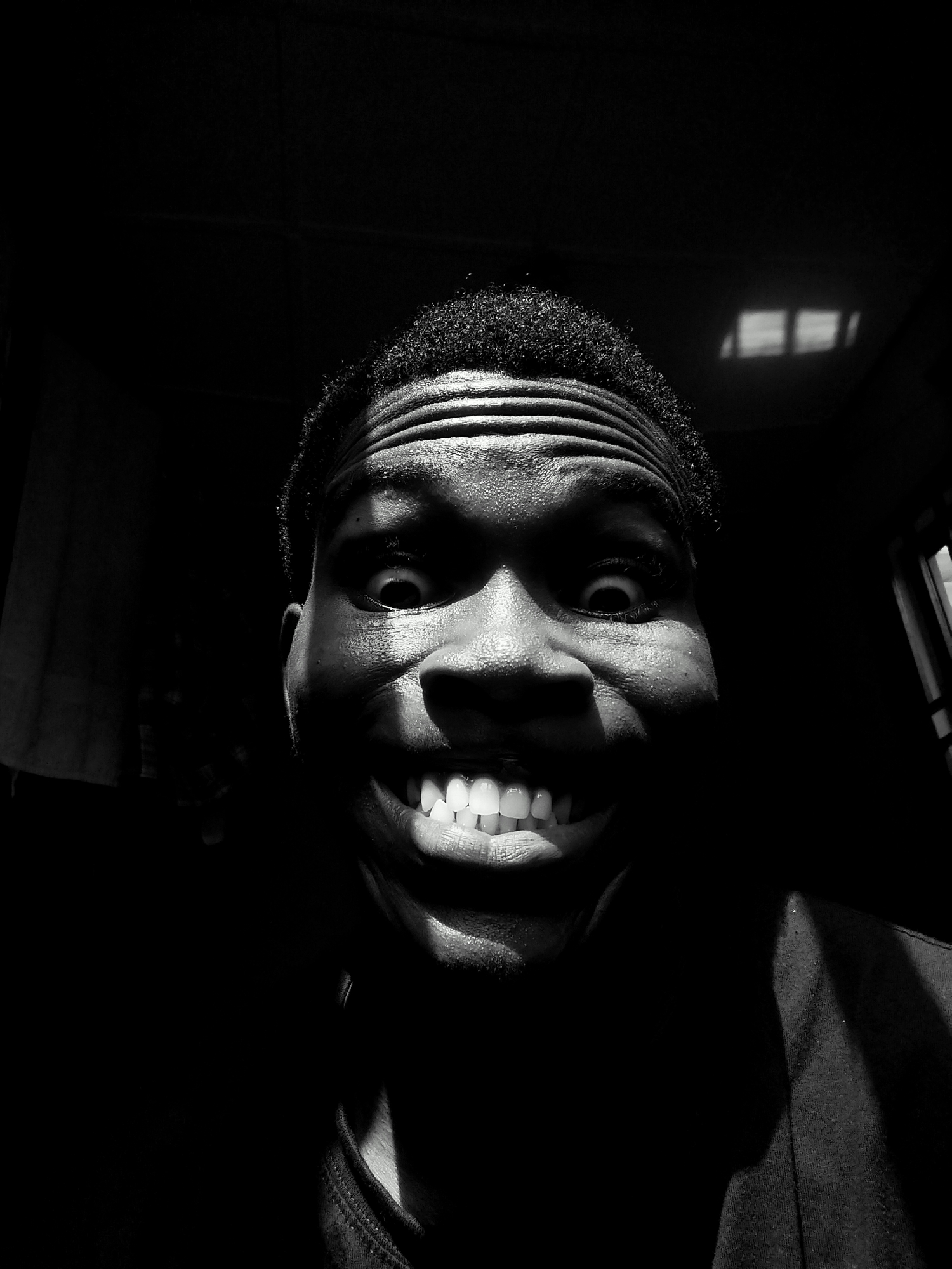 A black and white close-up photograph depicts a person with dark skin grinning widely. The person's face is partially illuminated, with the upper portion, including the forehead and eyes, being more brightly lit than the lower portion. The background is mostly dark, but what appears to be the reflection of a window is visible on the wall behind the subject. The person is wearing a dark shirt, the collar of which is visible. There is no information in the image to verify that the location is Kubwa, Nigeria. The image features a dark, high contrast, and dramatic lighting effect.