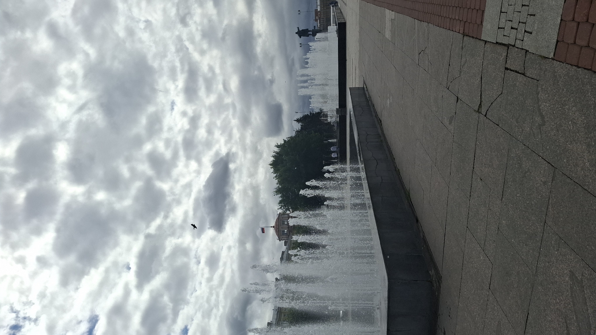 The image depicts a public square with multiple fountains spraying water. The sky is overcast with dramatic clouds. In the background, a building with a dome and the Russian flag is visible, along with a statue of a man with outstretched arms. Trees line the perimeter of the square. Paved walkways and a brick border are present in the foreground. The scene appears to be in Saint Petersburg, Russia, likely near the Narva Triumphal Arch or a similar public space. There are a few people visible in the distance, mostly walking or sitting. The fountains are the dominant feature, creating a dynamic visual effect with the water jets.