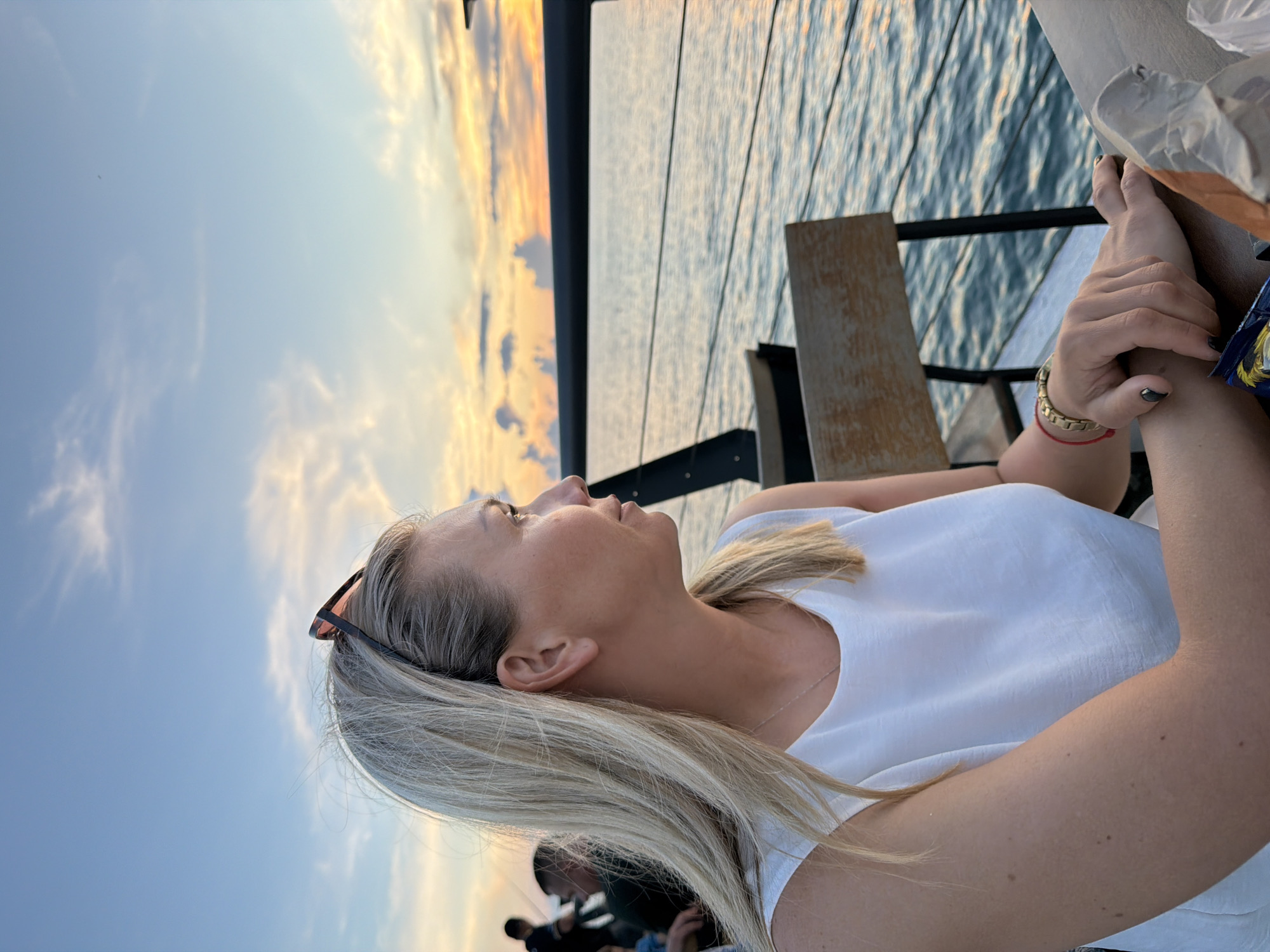 An outdoor, close-up, profile shot captures a Caucasian woman with blonde hair, wearing a white sleeveless top and sunglasses pushed back on her head, as she looks out at a calm body of water during sunset. The woman is seated at a table, with her hands resting on it. In the background, the setting sun casts a warm glow on the sky, with scattered clouds. The water is rippling, and a railing with chairs is visible along the waterfront. The overall scene suggests a relaxed evening setting at a seaside location.