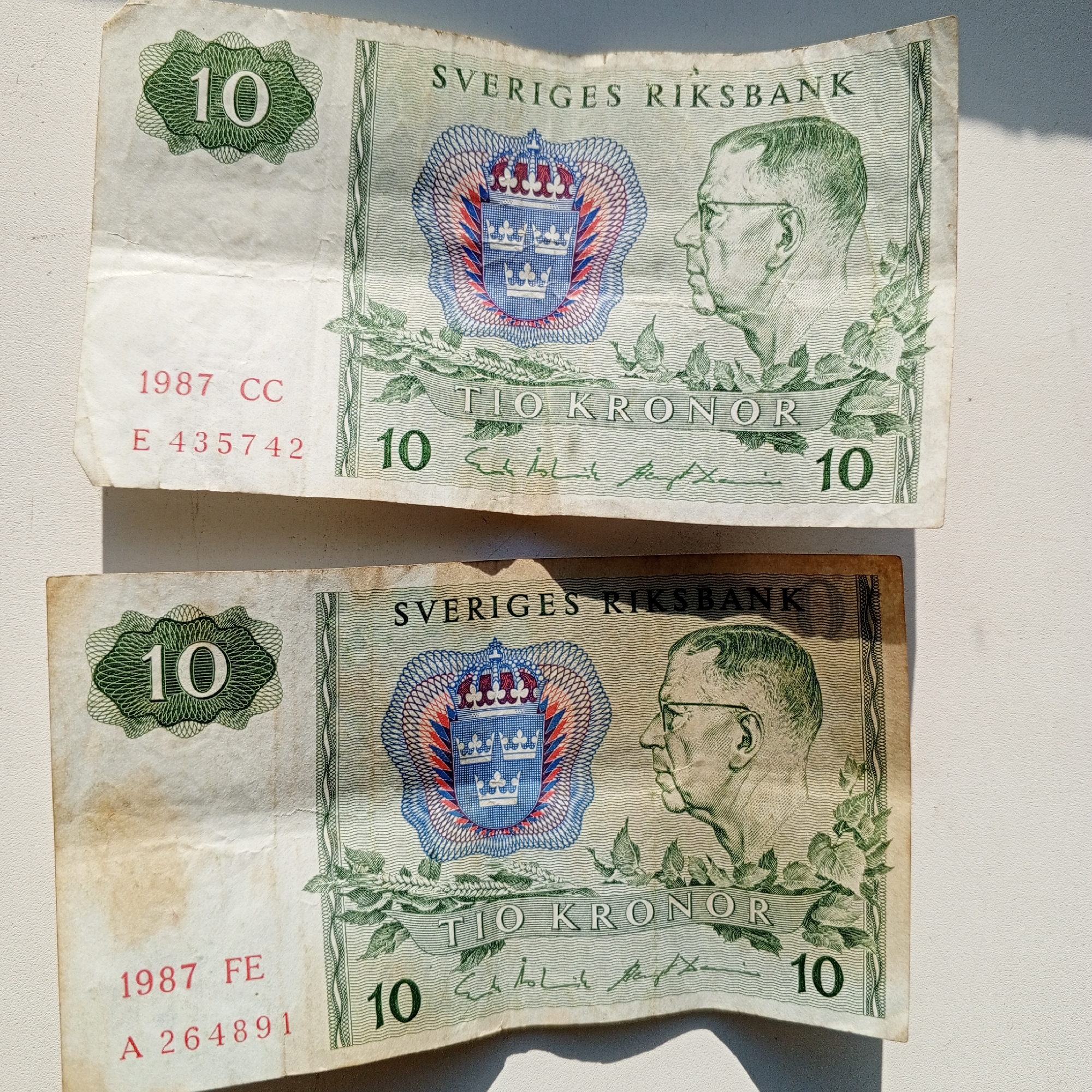 The image displays two 10 Swedish Kronor banknotes from 1987. Both banknotes feature a portrait of the Swedish king Gustav VI Adolf on the right, the Swedish coat of arms on the left, and the denomination "10" prominently displayed. The text "SVERIGES RIKSBANK" is visible at the top of both notes, indicating the issuing bank, and "TIO KRONOR" signifies "Ten Kronor."
The top banknote has the serial number "E 435742" and the date "1987 CC" stamped in red. The bottom banknote has the serial number "A 264891" and the date "1987 FE" stamped in red. The banknotes appear to be creased and slightly worn. The background is a plain, textured surface, possibly a table or countertop, illuminated by natural light casting a shadow. The banknotes are not located in Sayanogorsk, Russia.
