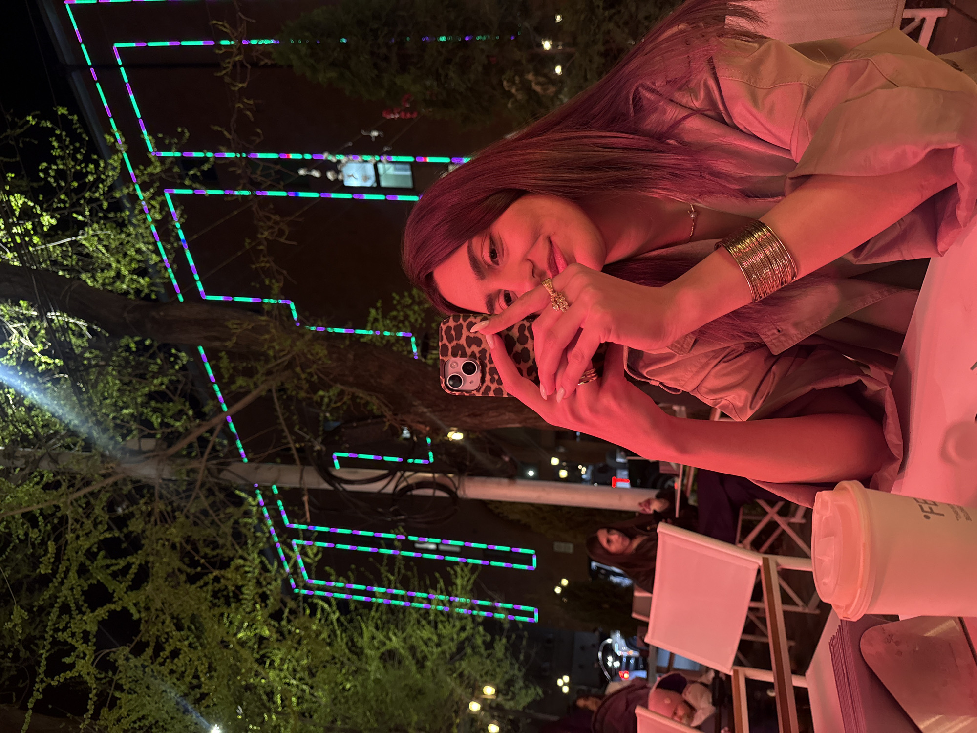 A woman holds a leopard-print smartphone, seemingly taking a photo or video, in Yerevan, Armenia. She is seated at an outdoor table. A white container rests on the table. In the background, trees are decorated with green and purple neon lights. Other people are visible in the background, seated at tables. It is nighttime.