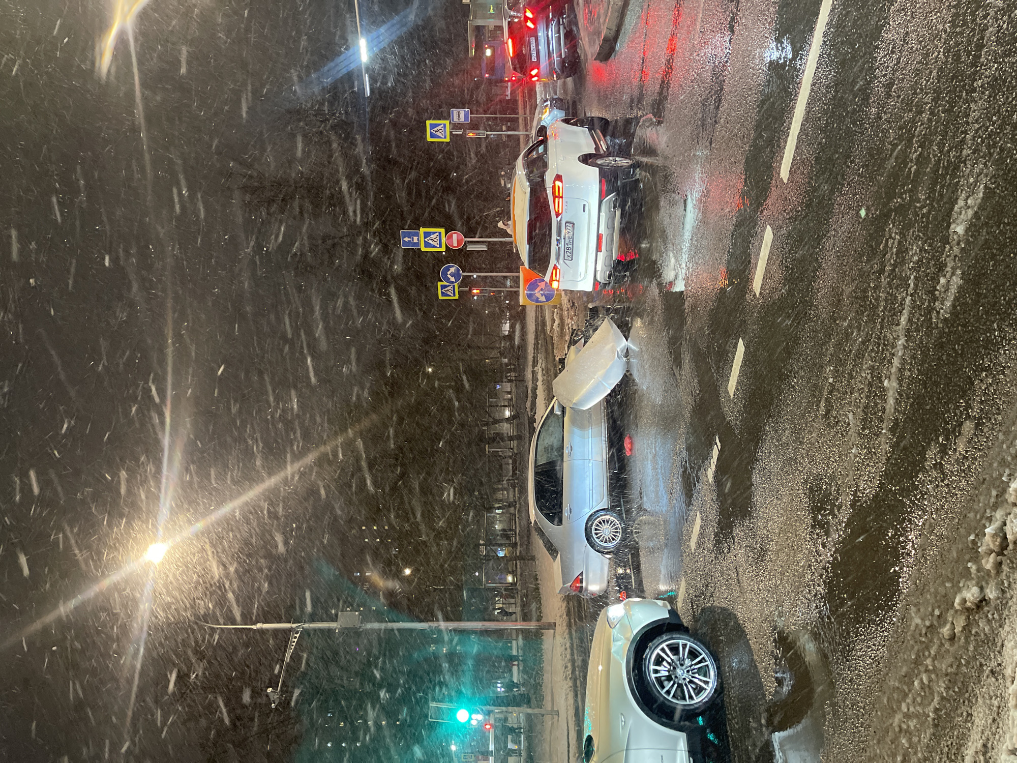 The image captures a nighttime scene on a city street in Moscow, Russia, during a heavy snowfall. The road surface is wet, slushy, and covered with patches of snow, reflecting the bright lights from streetlights and vehicles.
The central focus is on what appears to be a traffic incident involving multiple cars. In the foreground, part of a light-colored car is visible. Further back, a silver sedan has its hood fully open, suggesting damage or mechanical trouble, and its rear bumper appears damaged. Behind this sedan, a white SUV with visible rear damage, possibly from a collision, is stopped. Its Russian license plate, "X281HE 977," indicates registration in the Moscow region, and "POLAR" is visible on its rear. Another darker car is stopped behind the white SUV, with its rear lights illuminated and a partially visible license plate, "X27XH 77."
A cluster of Russian road signs, including multiple pedestrian crossing signs, a 'no entry' sign, and a 'give way' sign, stands on the right side of the road. Traffic lights are visible, with green lights in the lower left of the frame and red lights further down the road. The street is illuminated by bright streetlights, which cause lens flares and highlight the falling snowflakes, creating visible streaks and dots across the dark sky. The heavy snow contributes to poor visibility and challenging driving conditions.