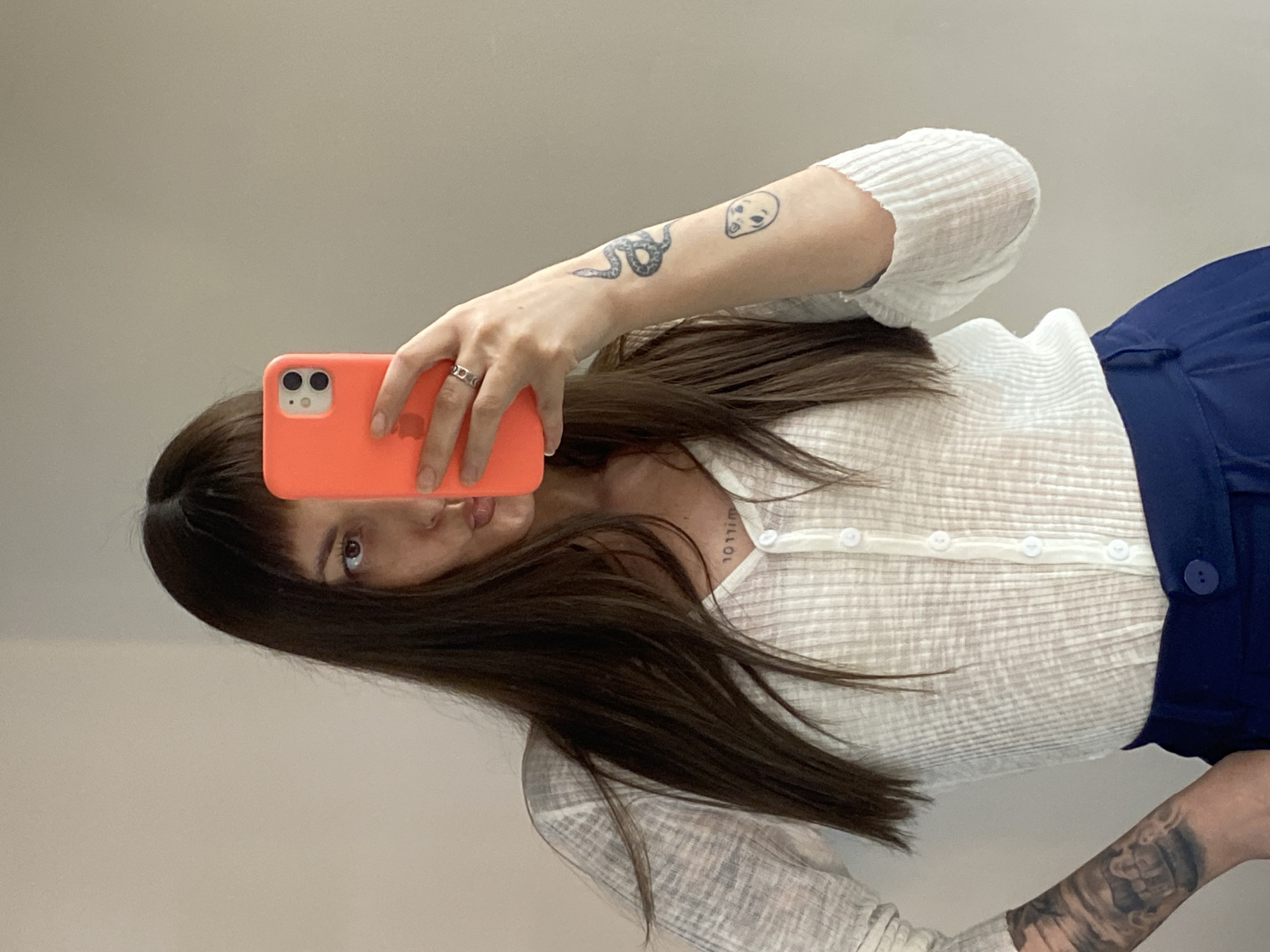The image shows a woman taking a selfie. She is holding an orange phone up to her face. She has brown hair and is wearing a white shirt and blue pants. Tattoos are visible on her left arm and neck. The background appears to be a plain white wall. It's impossible to determine whether this image was taken in Votkinsk, Russia.