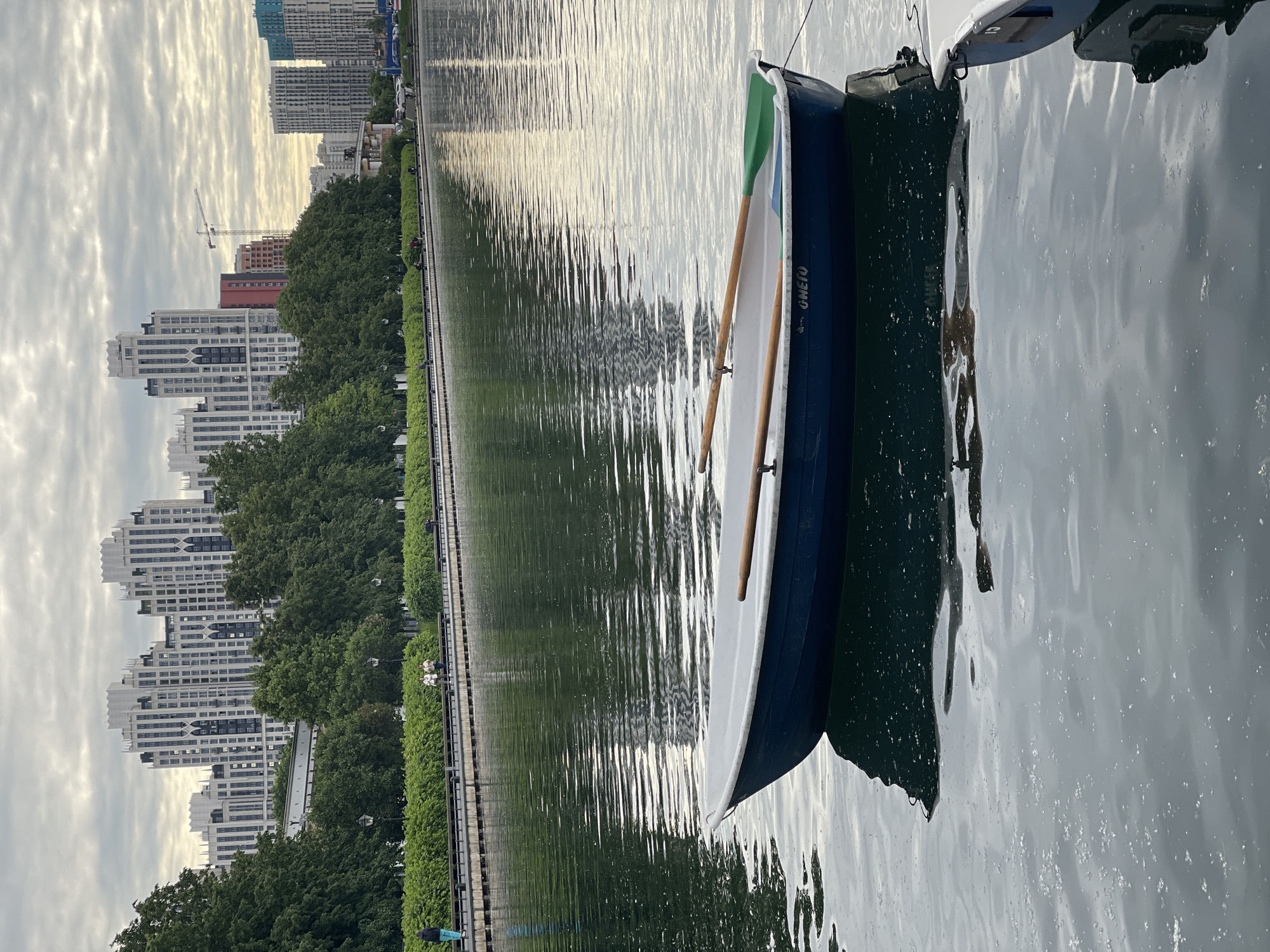 An urban waterscape is depicted under a partly cloudy sky, with a calm body of water in the foreground and midground, and a city skyline in the background. A small boat, possibly a rowboat or dinghy, is moored on the right side of the frame. The boat is dark blue on the hull and white on the interior, with two wooden oars resting inside, one showing a green tip. The word "GWETO" is visible in white lettering on the blue side of the boat. Another white object, likely part of another boat or a dock, is partially visible next to it.
The water reflects the light from the sky and distant elements, creating shimmering patterns. Along the opposite bank, a green treeline borders a pathway or promenade. Beyond the trees, numerous modern high-rise buildings form a city skyline. These buildings vary in color and design, with some appearing light blue, white, or reddish-brown, and one distinctive white building featuring a stepped roofline. A construction crane is visible in the distance, suggesting ongoing development. A few indistinct figures of people can be seen on the pathway in the distance. The soft light and reflections suggest either early morning or late afternoon, contributing to a peaceful atmosphere.