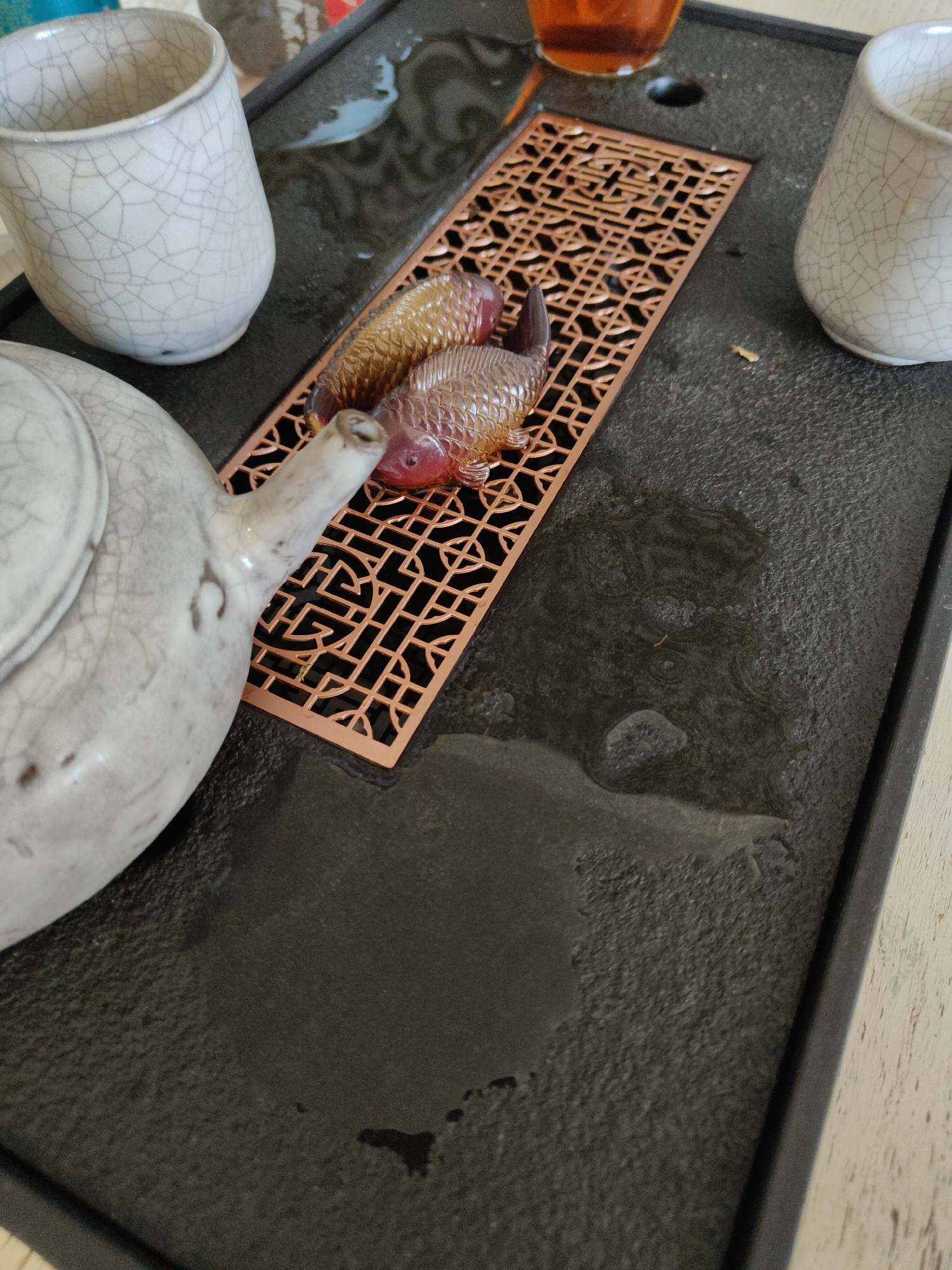 The image captures a tea ceremony setup on a black tray. A white teapot is positioned on the left side, with its spout pointing towards the center. Two teacups with a crackled glaze finish are placed on either side of the tray. A decorative bronze-colored, rectangular lattice sits in the middle, adorned with two ornamental fish figurines. The tray's surface is wet, reflecting light and suggesting recent use. The scene is likely set indoors, but the exact location and city are unknown.