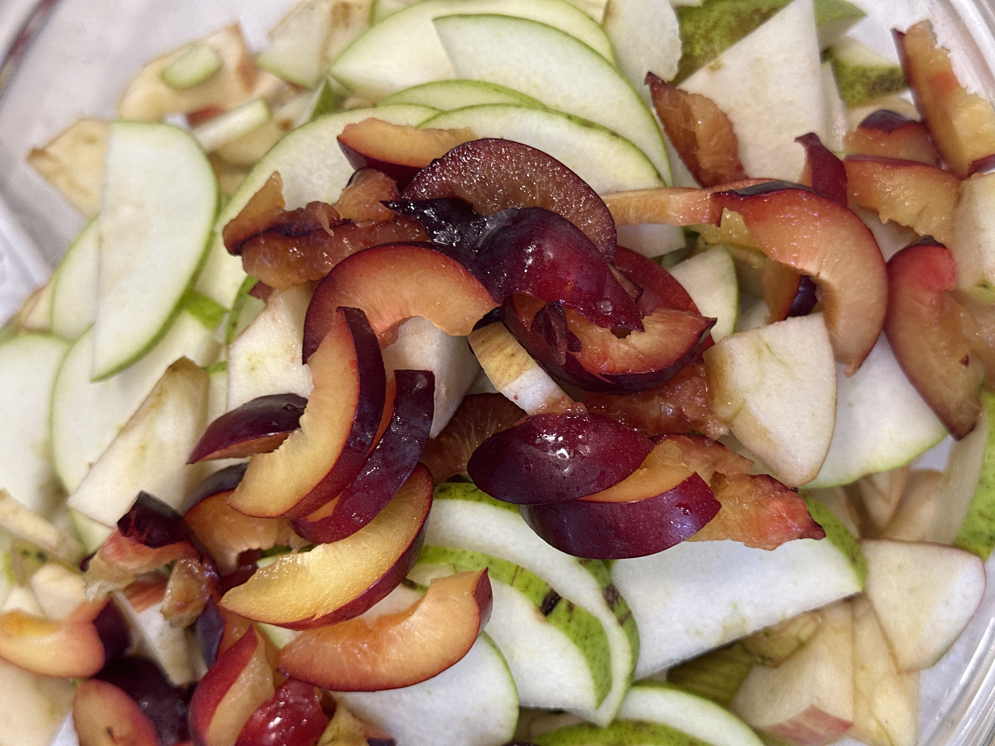 A close-up, overhead shot showcases a medley of sliced fruits, predominantly apples and plums, arranged in a clear plastic container. The apples are cut into thin, crescent-shaped slices, revealing their pale yellow flesh and green or reddish skin. Interspersed among the apple slices are vibrant plum pieces, displaying a spectrum of colors from deep purple to a rosy pink and orange hue. Some plum slices are cut into wedges, while others are smaller, more fragmented pieces. The fruits appear fresh and possibly prepared for a salad or dessert. The background is indistinct, focusing entirely on the fruit. There is no indication of the city of Nevinnomyssk, Russia, or any other specific location within the image.