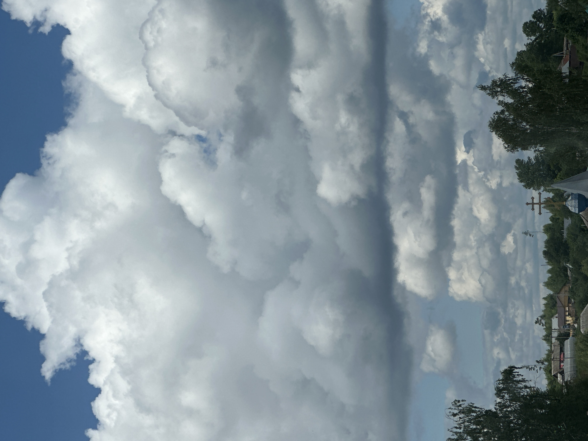 The image shows a cloudy sky with patches of blue visible. The clouds are white and gray, suggesting varying densities. The ground, visible on the right side of the image, is populated with trees and buildings of an unidentified city. No people are visible. A cross is observed atop the roof of an unknown structure. The scene depicts an outdoor environment under a partly cloudy sky.