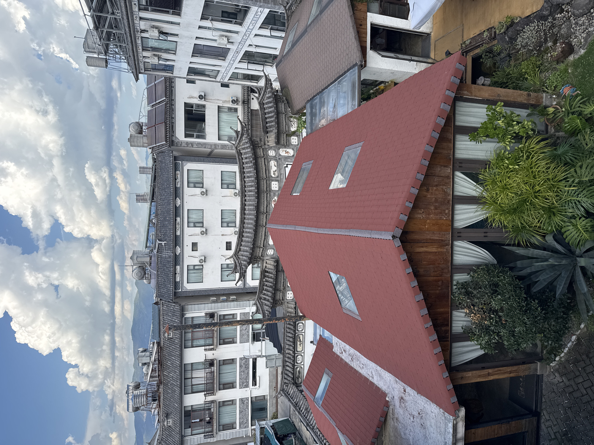 This is an aerial view of rooftops in Dali City, China. The foreground features a building with a red, segmented roof, incorporating diamond-shaped skylights. This building appears to be a restaurant or cafe, with large windows, white curtains, and lush green plants visible in front. Behind this structure, traditional Chinese-style tiled roofs are visible, adorned with decorative eaves. Further in the background, modern multi-story buildings with white facades, balconies, and air conditioning units are prominent. Some of these buildings have water heaters and solar panels on their roofs. In the distance, a mountain range is visible under a sky filled with white, fluffy clouds against a blue backdrop. The overall scene depicts a mix of traditional and modern architecture within a scenic mountainous setting.