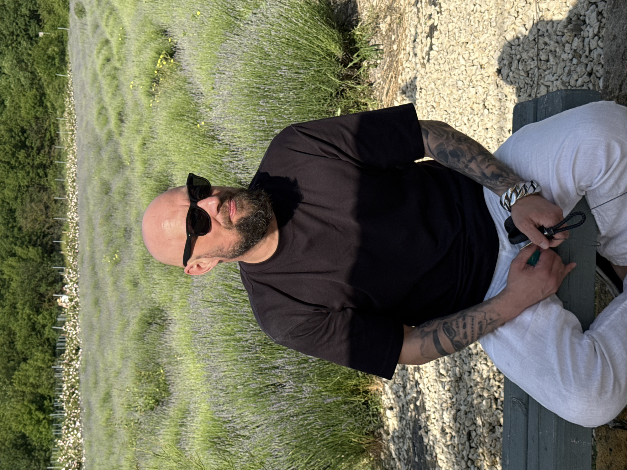 A bald, bearded man wearing sunglasses and a black t-shirt sits on a green bench in front of a lavender field. The man is positioned in the center of the frame, with his body angled slightly to the right. His left arm, adorned with tattoos, rests on his lap, while his right hand holds a green object. The background features a vast expanse of purple lavender flowers stretching towards a line of trees and vegetation. The ground in the foreground is covered with small, light-colored stones. The lighting suggests it is daytime, with sunlight casting shadows.