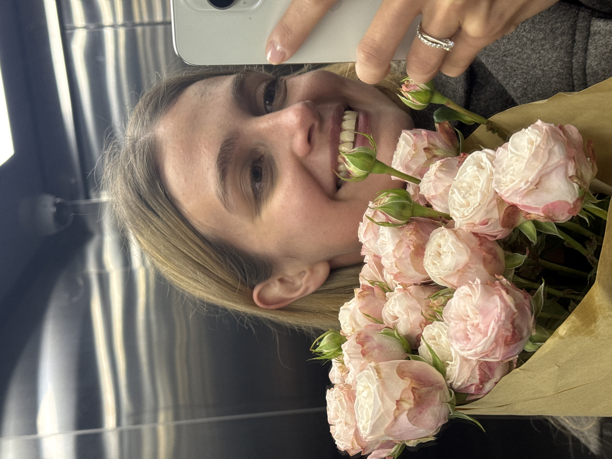 A woman with blonde hair is holding a bouquet of pink and white roses while taking a selfie in a mirrored elevator. She is wearing a gray coat and a diamond ring on her right ring finger. The background reflects the elevator's interior, with its metallic walls and lighting. The location is unspecified.
