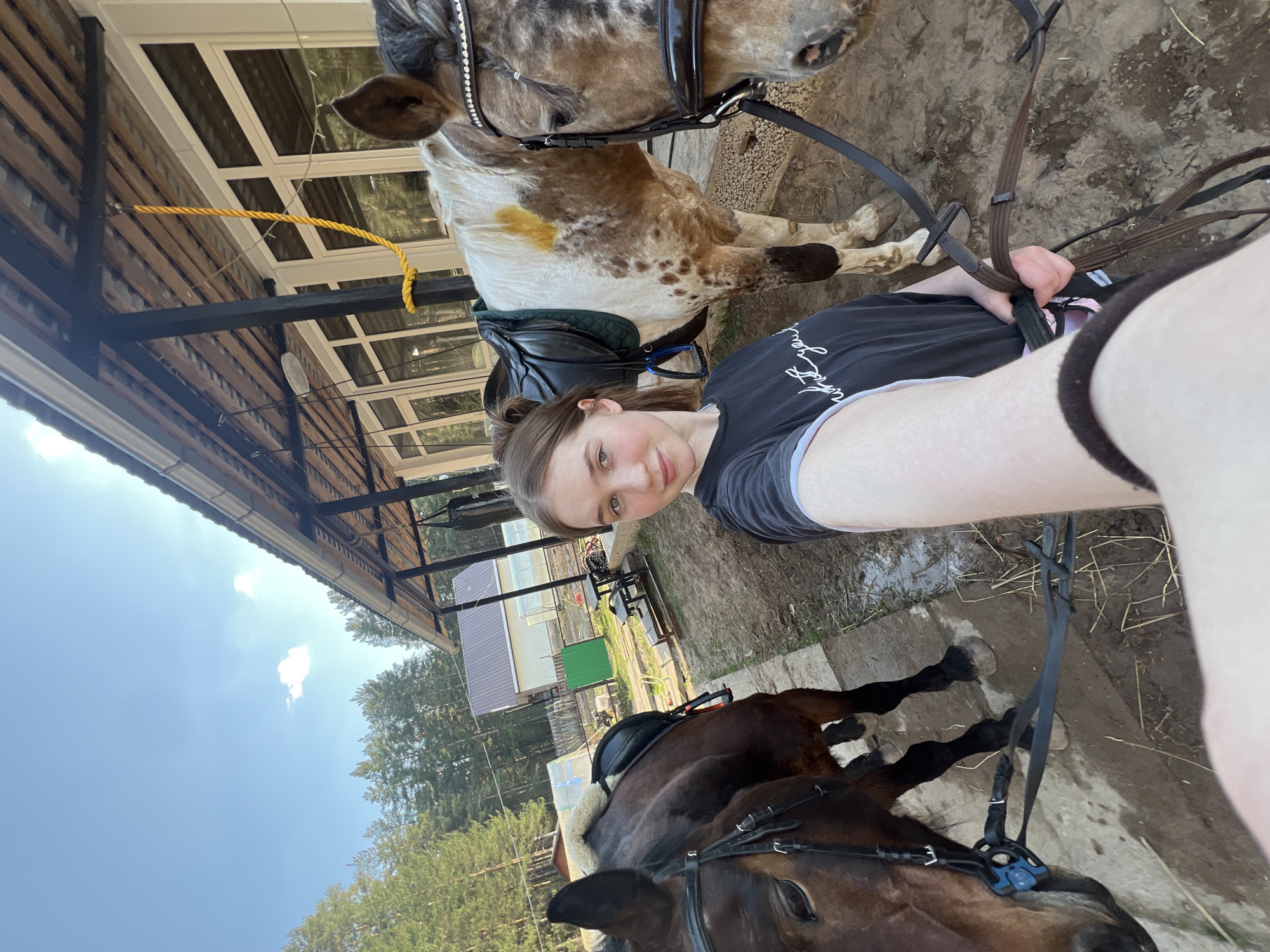 A young woman is taking a selfie with two horses in Chaikovsky, Russia. The photo appears to be taken outdoors, near a stable or horse riding facility. The woman is holding the reins of the horses. One horse is a light brown and white Appaloosa and is wearing a black saddle. The other is a dark brown horse, also wearing a saddle. The environment includes a wooden building with windows, a rope tied to an overhead beam, trees, and a blue sky with clouds.