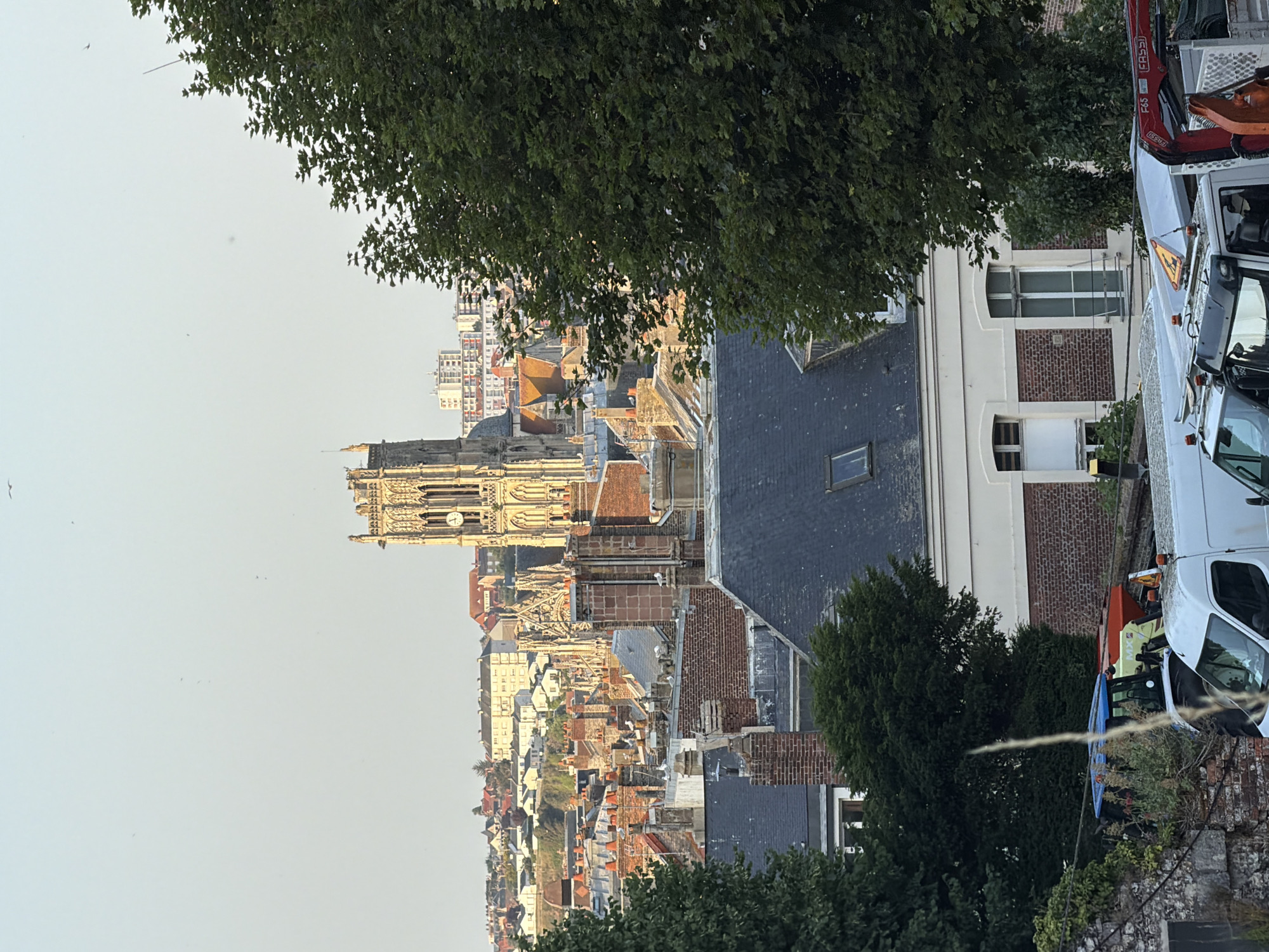 This image captures a view of the city of Dieppe, France, dominated by the prominent structure of the Saint-Jacques Church in the background. The church, with its tall, ornate bell tower, stands out against the sky, its golden stone illuminated by the setting sun. In the foreground, the rooftops of Dieppe are visible, a mix of red-tiled and grey slate surfaces. Several brick chimneys punctuate the rooflines, some adorned with white seagulls perched on top.
The middle ground reveals more of the urban landscape, with buildings of varying architectural styles, some featuring red-tiled roofs and others with modern facades. Further in the distance, a cluster of taller, more contemporary buildings can be seen. The foreground is partially obscured by the lush green foliage of trees, framing the scene. Below the rooftops, in the lower part of the image, the top of a white van is visible, its roof equipped with orange warning lights. Next to it, the cabin of a blue tractor is partially visible, along with a red piece of machinery, possibly a crane, attached to another vehicle. The overall impression is of a city scene at dusk, with the silhouette of the church serving as a focal point.