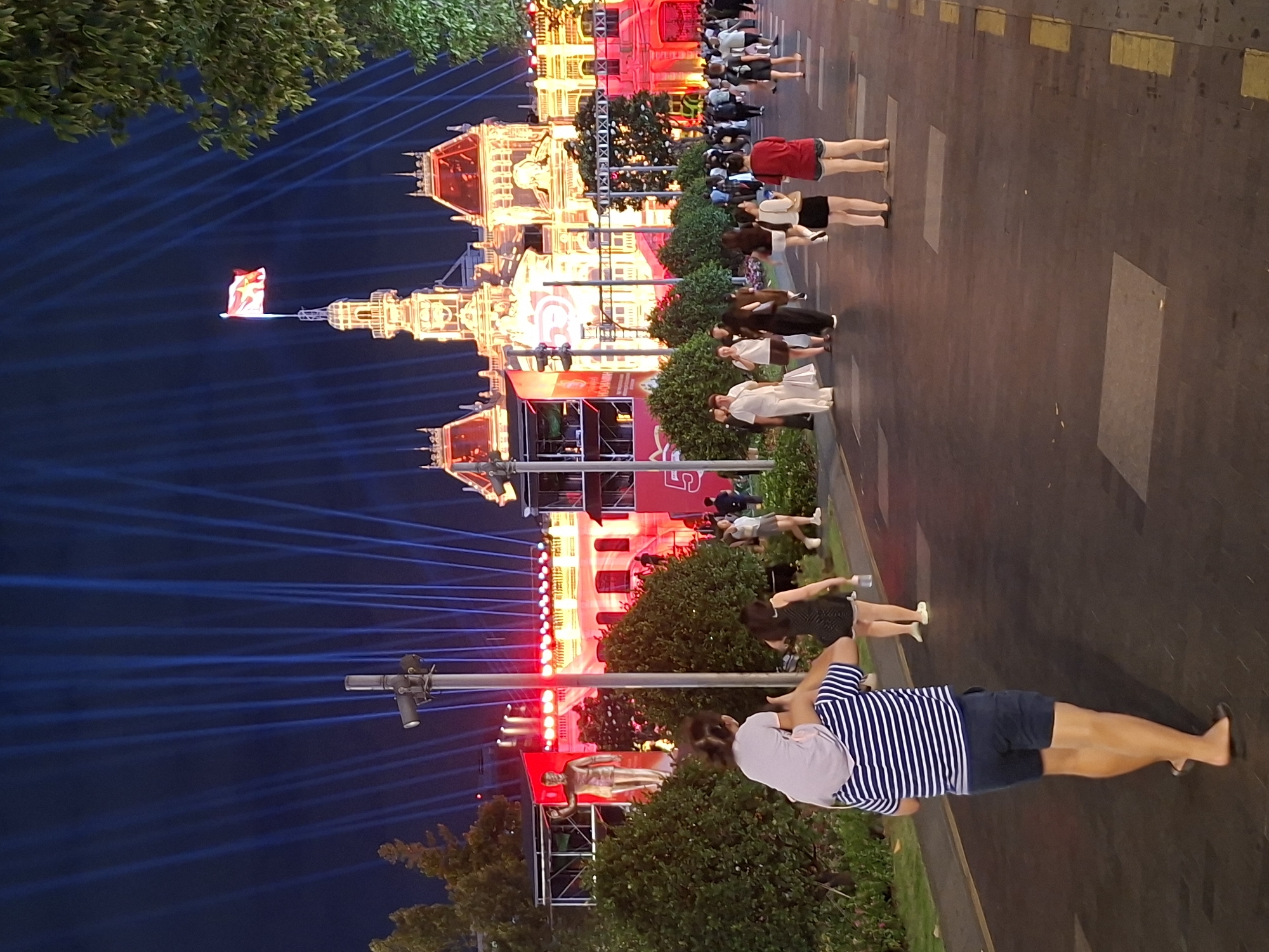 The image depicts a nighttime scene in Thủ Đức, Vietnam, likely during a public event or celebration. In the foreground, a man wearing a blue and white striped t-shirt and navy shorts walks away from the camera, carrying a young child on his shoulders. The child is wearing a light-colored shirt. Behind them, other individuals are walking along a paved pathway.
The background features a grand building illuminated with warm yellow and red lights, suggesting it is a prominent landmark. A flag flies from a tower atop the building. A series of bright blue light beams are projected upwards into the night sky from behind the building, creating a dramatic effect. Lush green bushes line the pathway in the middle ground. The overall atmosphere appears festive and lively.
