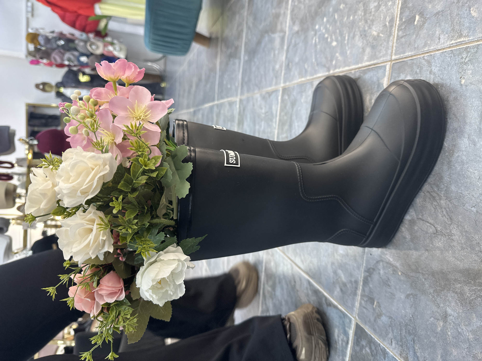 The image depicts a pair of black rain boots filled with a bouquet of pink and white flowers. The boots are positioned on a gray tiled floor. In the background, there are indistinct figures and items, possibly within a store setting. A glimpse of a person wearing dark clothing and brown shoes is visible at the bottom of the image. There is no discernible evidence in the image to definitively associate it with the city of Chaikovsky, Russia.