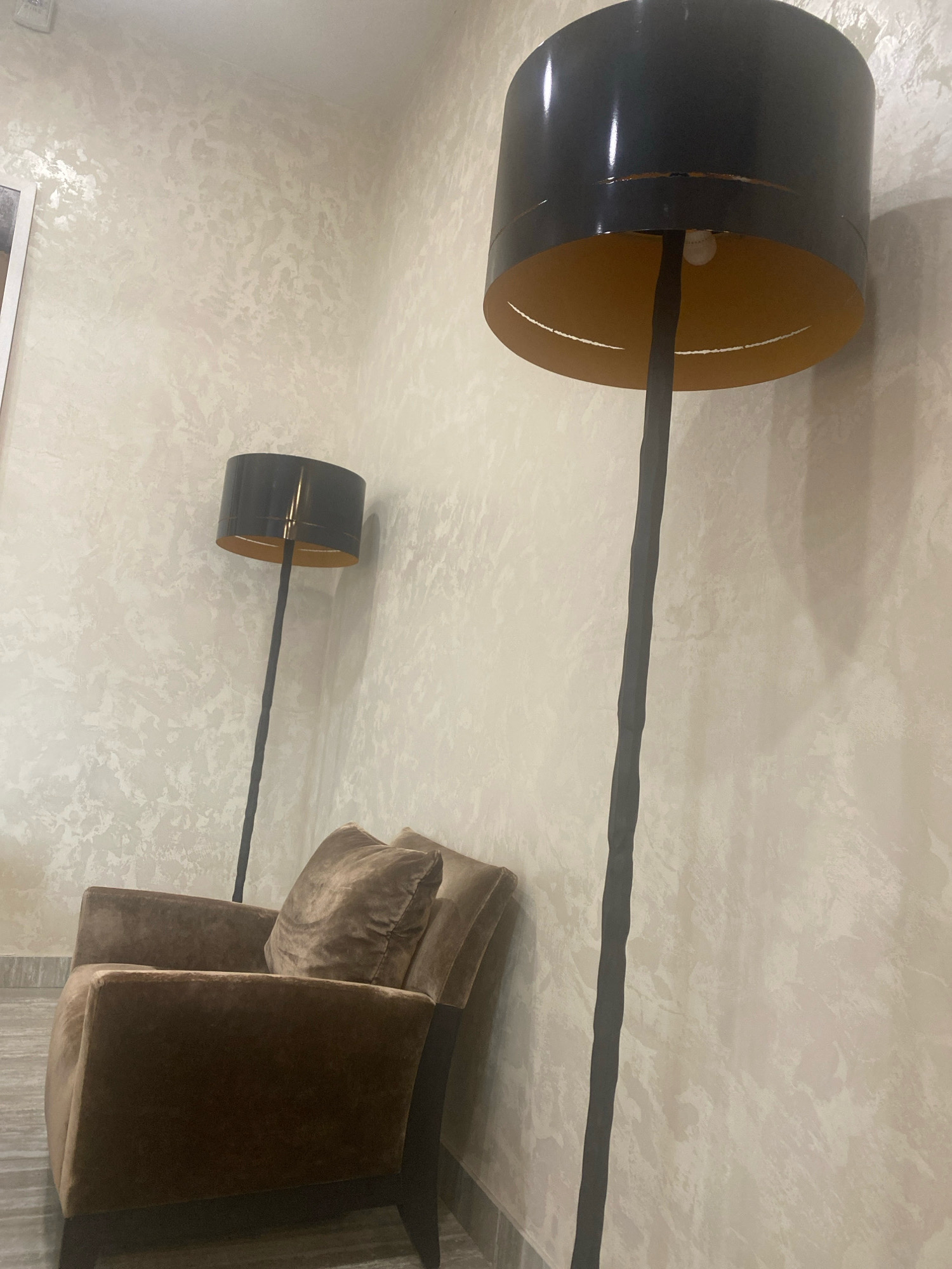 The image depicts an indoor scene, likely a living room or lounge area. In the foreground, a plush brown armchair is positioned on a tiled floor. Behind the armchair, two tall floor lamps with black cylindrical shades and gold interiors are visible. The walls are textured and light-colored, with a subtle pattern. The lighting suggests it might be evening or a dimly lit space. There are no people visible in the image. The scene is static and does not suggest any specific activity. The location is stated to be in the city of Minsk, Belarus.