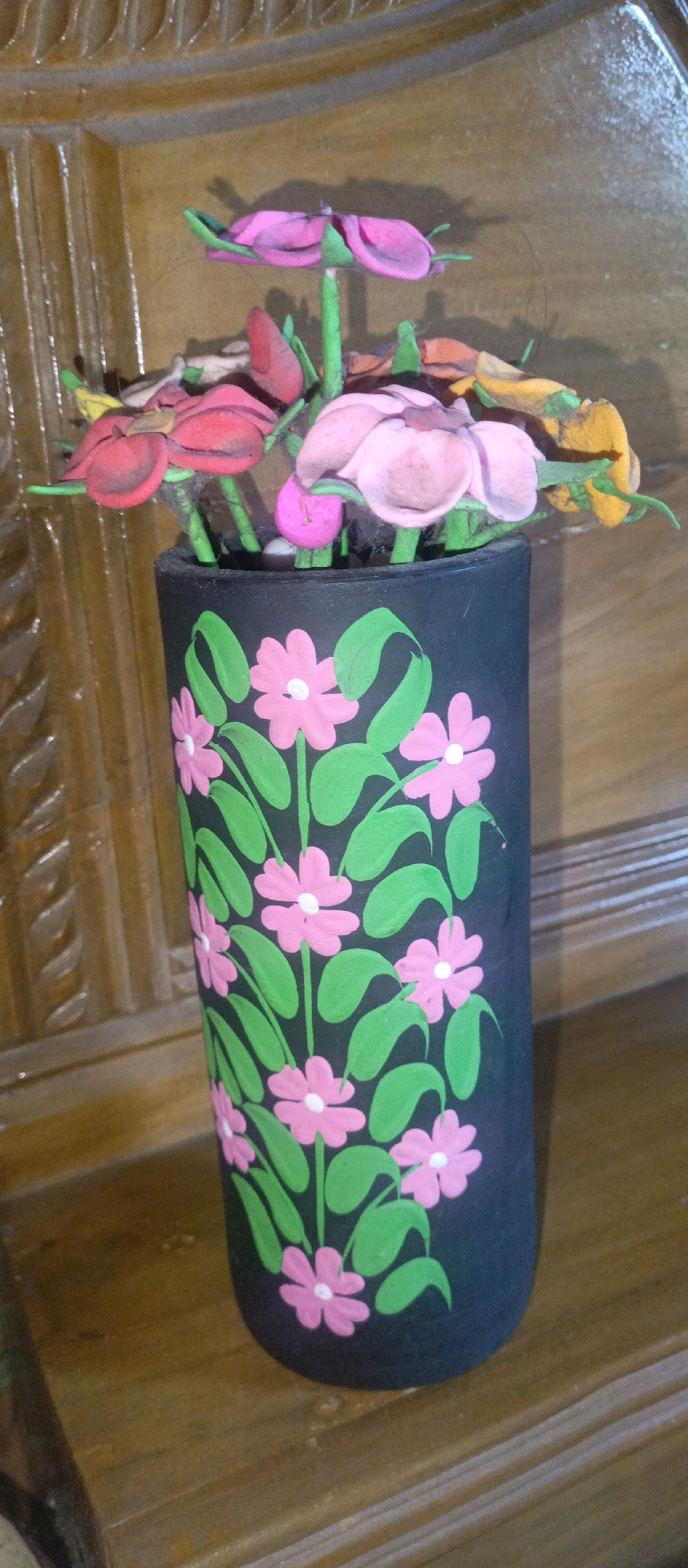A close-up, slightly tilted, eye-level shot shows a black cylindrical vase decorated with painted pink flowers and green leaves. The vase is filled with various handmade clay flowers in colors of pink, red, yellow, and orange, with green stems. The vase is positioned on a wooden surface that appears to be a table or shelf. The background is a textured wooden surface with vertical grooves and some carved detailing at the top, suggesting it is furniture, possibly a cabinet or part of a room's decor. The lighting is moderate, casting some shadows. The scene appears to be indoors. Without further context, specific location details in Gazipur, Bangladesh, cannot be determined from the visual information provided.