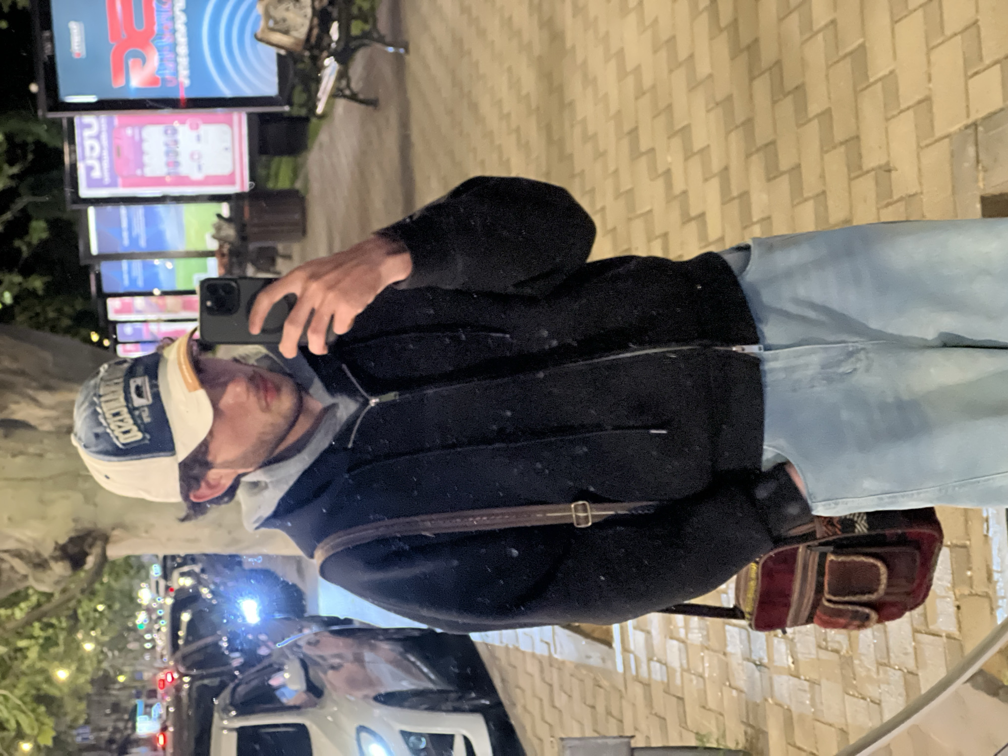 A person is holding a phone and taking a picture in Yerevan, Armenia. The person is wearing a cap, a gray hoodie, and a black jacket with a brown strap. They are also wearing light blue jeans and black gloves. There is a maroon bag. The background features street lights, traffic, and digital billboards showing advertisements. The street is paved with cobblestones.