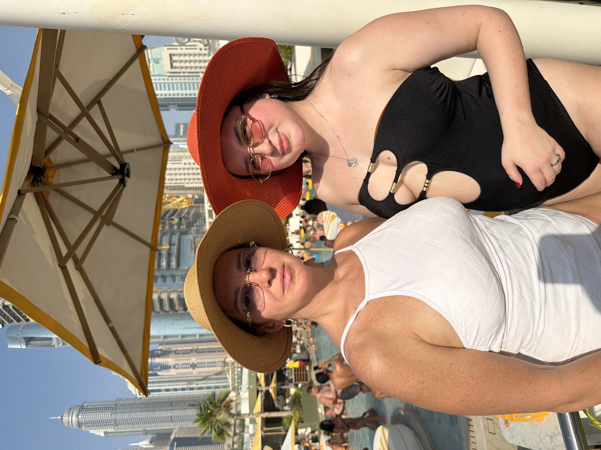 Two women stand at a poolside in Dubai, United Arab Emirates, under a large umbrella with a yellow trim. In the background, the city skyline is visible, featuring numerous skyscrapers. The woman on the left wears a white swimsuit, a straw hat, and sunglasses. The woman on the right wears a black cutout swimsuit, an orange hat, and sunglasses. Both women are looking towards the camera. Other people are visible in the pool area, swimming and relaxing. The scene appears to be a sunny day, likely at a resort or hotel.