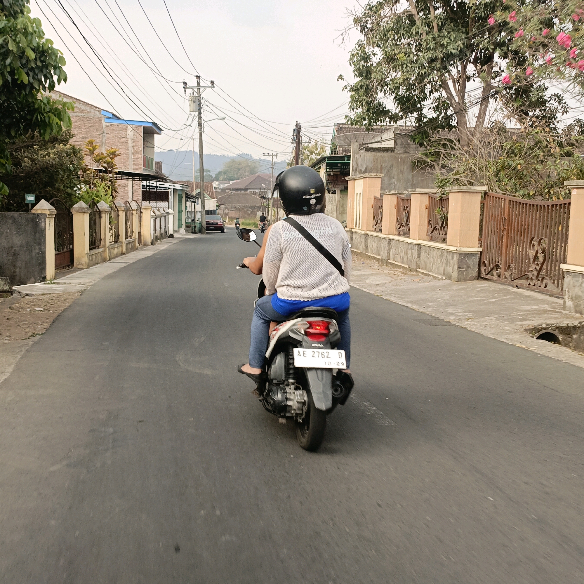 This is a rear view, ground-level, medium shot of two people riding a scooter down a paved street in Salatiga, Indonesia. The person in the front wears a black helmet and a light-colored, woven sweater over a white shirt. The person in the back wears blue pants and black sandals. The scooter's license plate reads "AE 2762 D". The street is lined with houses and shops with beige fences and tiled roofs. Trees with green leaves and some pink flowers are visible. Utility poles and a dense network of overhead wires crisscross the sky, which is overcast and pale. The road surface is gray asphalt with some visible cracks and debris. The overall scene depicts a typical street scene in a residential or commercial area in Indonesia.