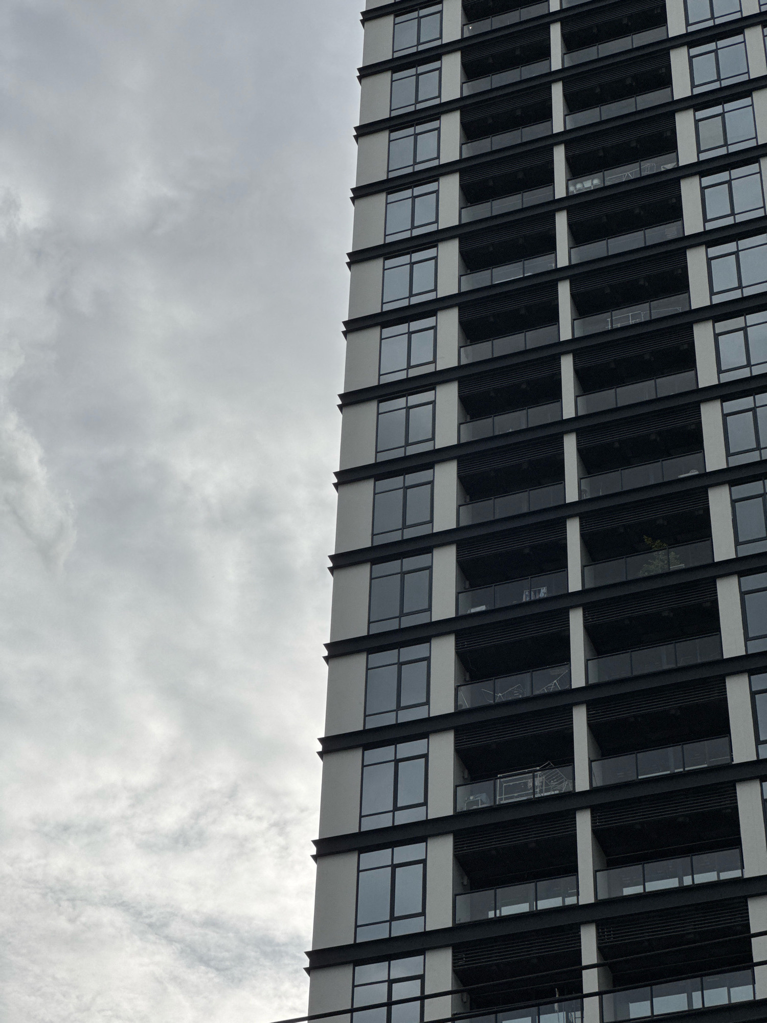 The image shows a close-up, low-angle shot of the side of a modern, multi-story apartment building. The building features a repeating pattern of windows and balconies. The windows are large and dark-tinted, set within light-colored frames. The balconies are recessed and have dark railings. The building's facade is predominantly light gray with black horizontal accents. The sky is overcast with light gray clouds. The composition is dominated by the building on the right side, with the sky occupying the left two-thirds of the frame. No people or specific identifiable objects are visible on the balconies or in the windows. The image appears to be taken during the daytime, given the illumination.