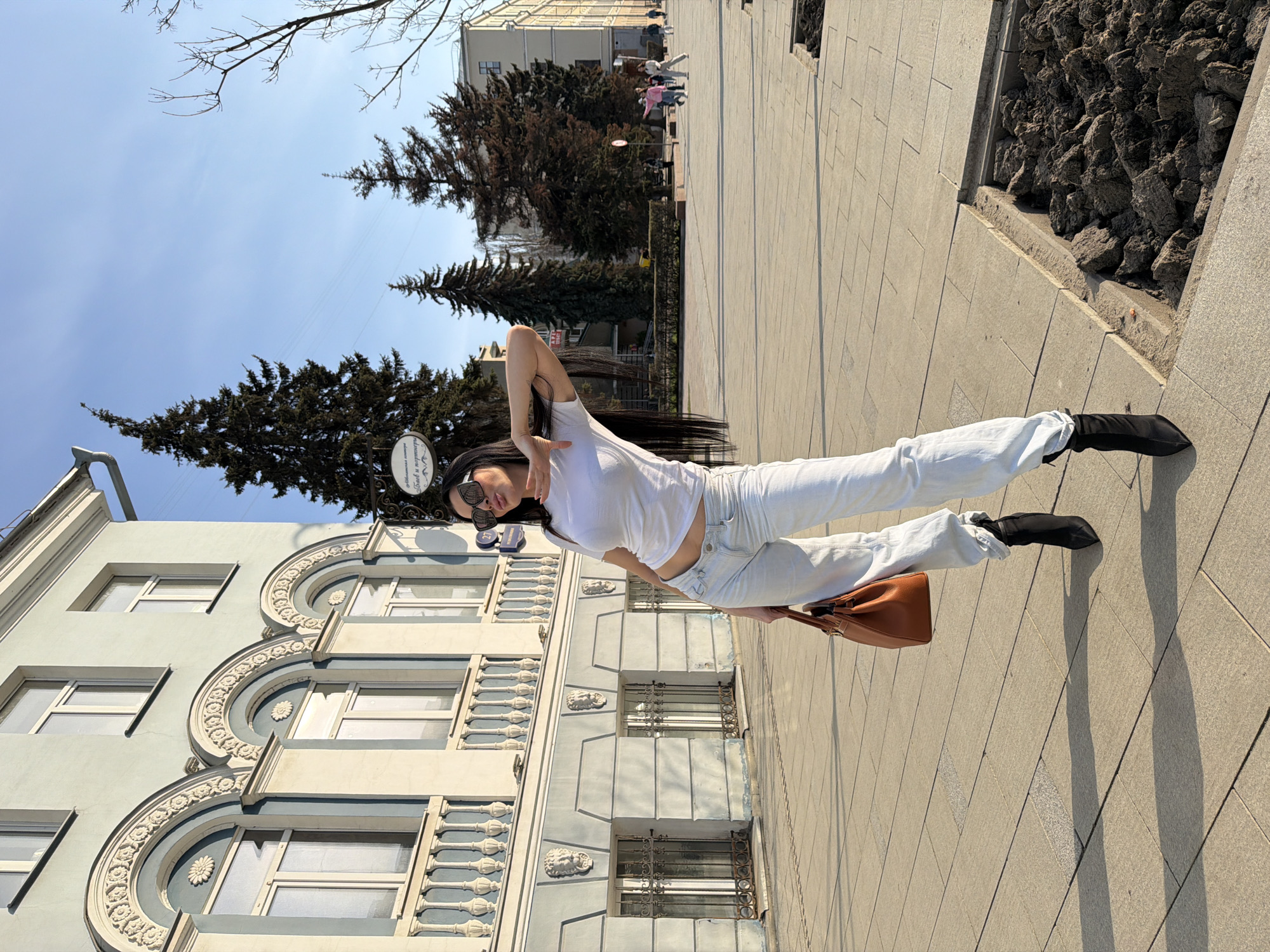 A woman wearing a white t-shirt, light blue jeans, and black shoes poses on a sidewalk in Voronezh, Russia. She holds a brown handbag and wears sunglasses. In the background, a building with ornate architectural details and arched windows is visible. Evergreen trees and a clear blue sky are also present. Other people are seen further down the sidewalk.