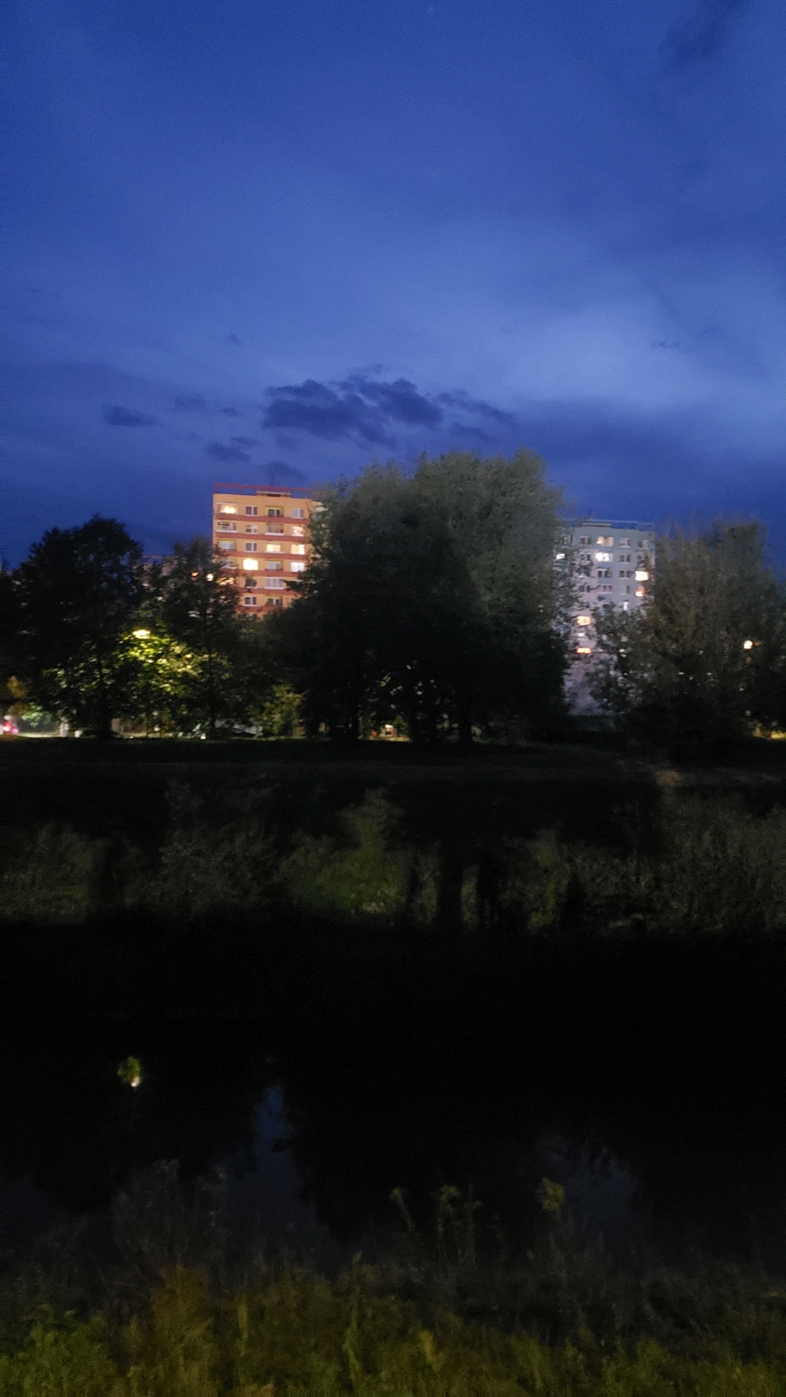The image captures a dusky twilight scene in Prešov, Slovakia, featuring a river in the foreground with its banks lined with dark vegetation and reeds. The river's surface reflects the sky and the silhouettes of trees. In the background, two apartment buildings are visible. The building on the left is a multi-story residential structure with an orange facade and illuminated windows, suggesting it is evening. To its right, another apartment building stands, this one with a lighter facade and also displaying lit windows. Trees are interspersed between the viewer and the buildings, with some illuminated by artificial light sources, casting a glow on the foliage. The sky above is a deep blue, transitioning into darker shades, with scattered clouds adding texture to the horizon.