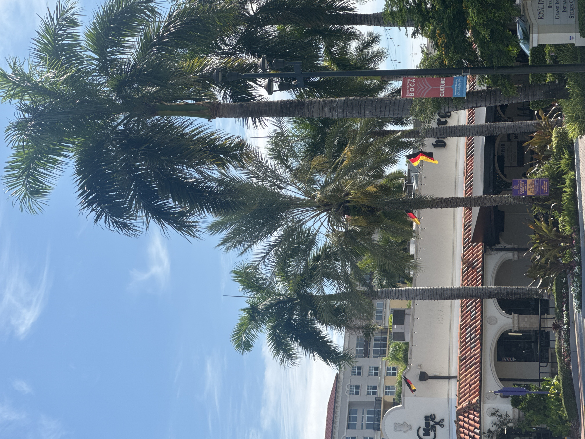 The image depicts a street scene in Boca Raton, Florida, United States of America, featuring a commercial building with Spanish-style architecture and a prominent tiled roof. Several tall palm trees are visible in the foreground, with their fronds reaching towards a bright blue sky with wispy clouds. The building has a white stucco facade with arched entrances and decorative details. A sign on the building reads "Man Cave." German flags are displayed on the building. A street lamp with multiple lights stands near a palm tree, and a banner on a lamppost advertises "Downtown Boca Culture." In the background, a multi-story building with balconies and numerous windows can be seen. The overall environment suggests a warm, sunny day in a developed urban area.