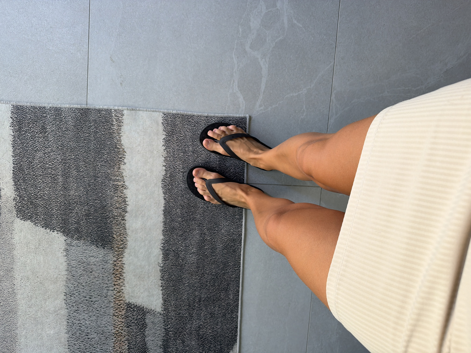A high-angle shot captures a person's legs from the knees down, clad in a light beige ribbed skirt and black flip-flops. The person is standing on a grey tiled floor, partially on a rug with a geometric pattern in shades of grey, white, and beige. The lighting is bright, casting subtle shadows. The image provides no specific information about the location within Finestrat, Spain, or any interactions.