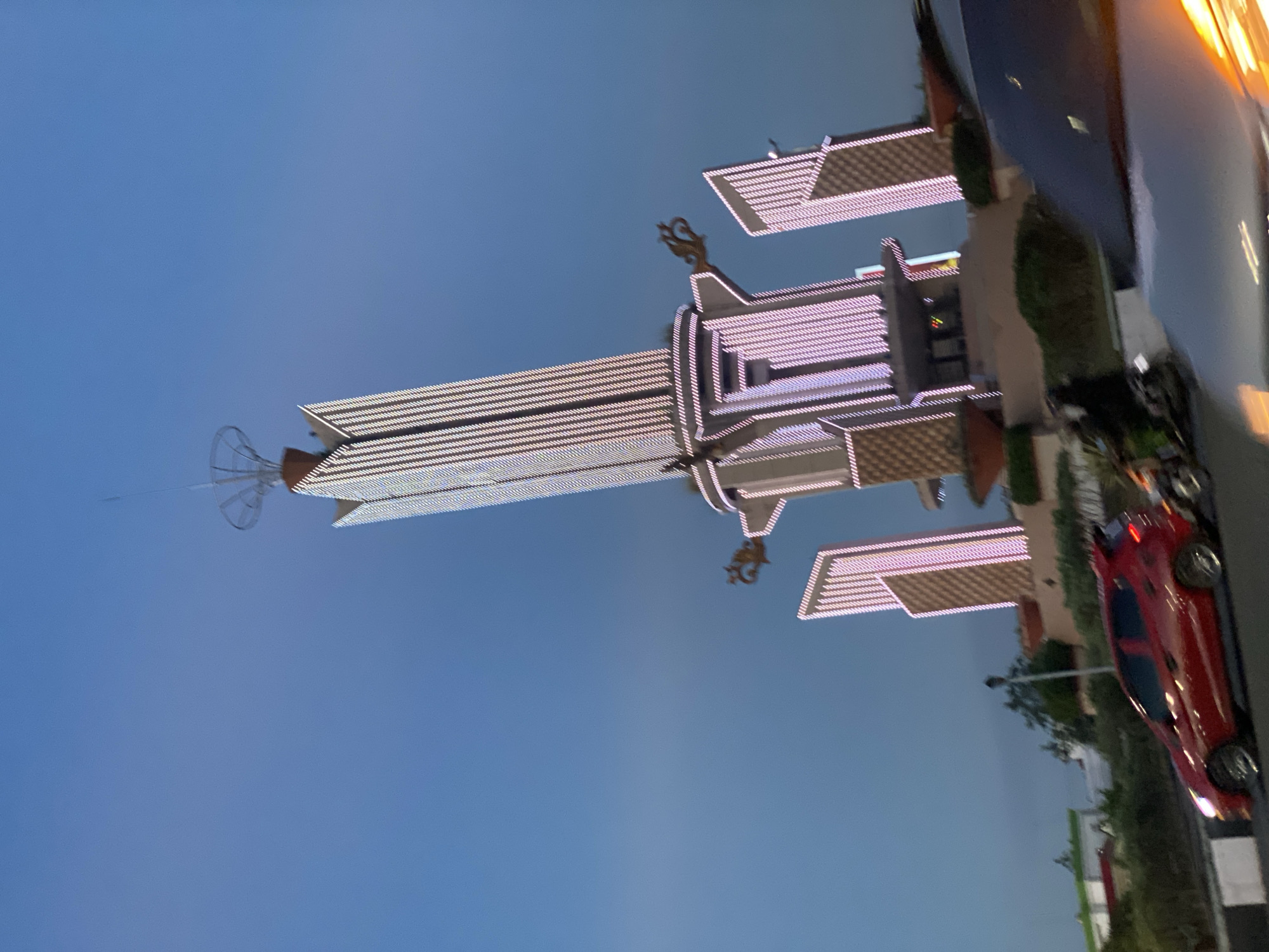 In Banjarbaru, Indonesia, at dusk, a tall, illuminated monument stands against a clear blue sky. The monument is adorned with numerous white and pink lights, creating a striking visual effect. Flanking the main structure are two smaller, rectangular towers, also lit with pink lights. Decorative golden elements are visible on the sides of the monument. In the foreground, a red car is driving on a road, with a motorcycle behind it. The road is bordered by white and black barriers. Green foliage and other structures can be seen in the background. The overall scene depicts a vibrant urban landscape at twilight.