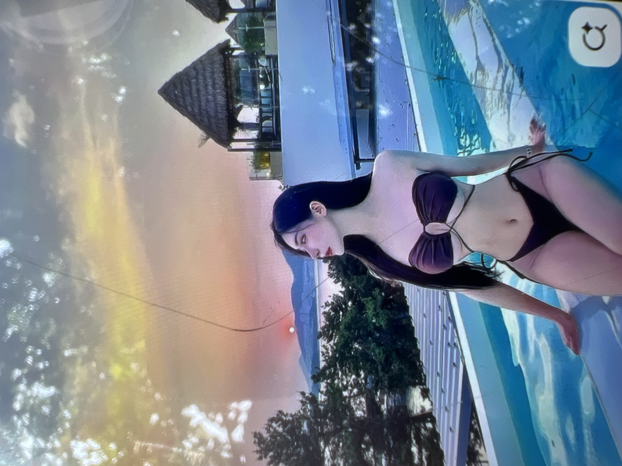The image depicts a young woman with long dark hair, wearing a dark bikini, posing by the edge of an infinity pool. The pool water is a bright blue, reflecting the sky and surrounding environment. In the background, there are thatched-roof structures, lush green trees, and a body of water with mountains in the distance. The sky is a gradient of orange and pink, suggesting it is either sunrise or sunset. The woman is looking to her left, with her left hand resting on the pool's edge. The overall scene suggests a tropical or resort-like setting.