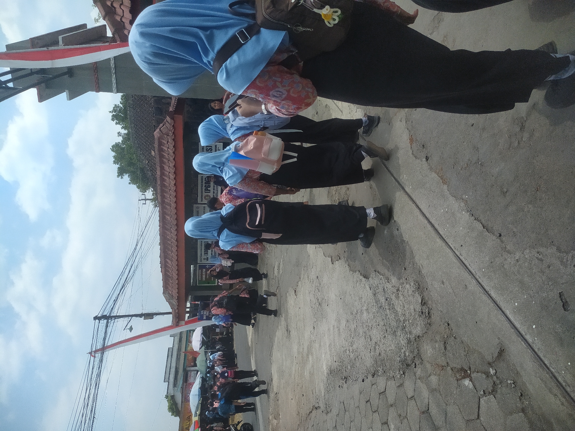 In the city of Candirejo, Indonesia, a group of students, most wearing blue hijabs and dark skirts, are walking on a paved road. The students are carrying backpacks and some are wearing patterned shirts beneath their hijabs. In the background, buildings with tiled roofs are visible, along with a large overhead structure decorated with red and white flags. The road is a mix of asphalt and paving stones, with some areas showing wear and tear. The sky is partly cloudy. Several students are captured mid-stride, suggesting movement and activity within the scene. The overall impression is of a school day or an event involving students outdoors.