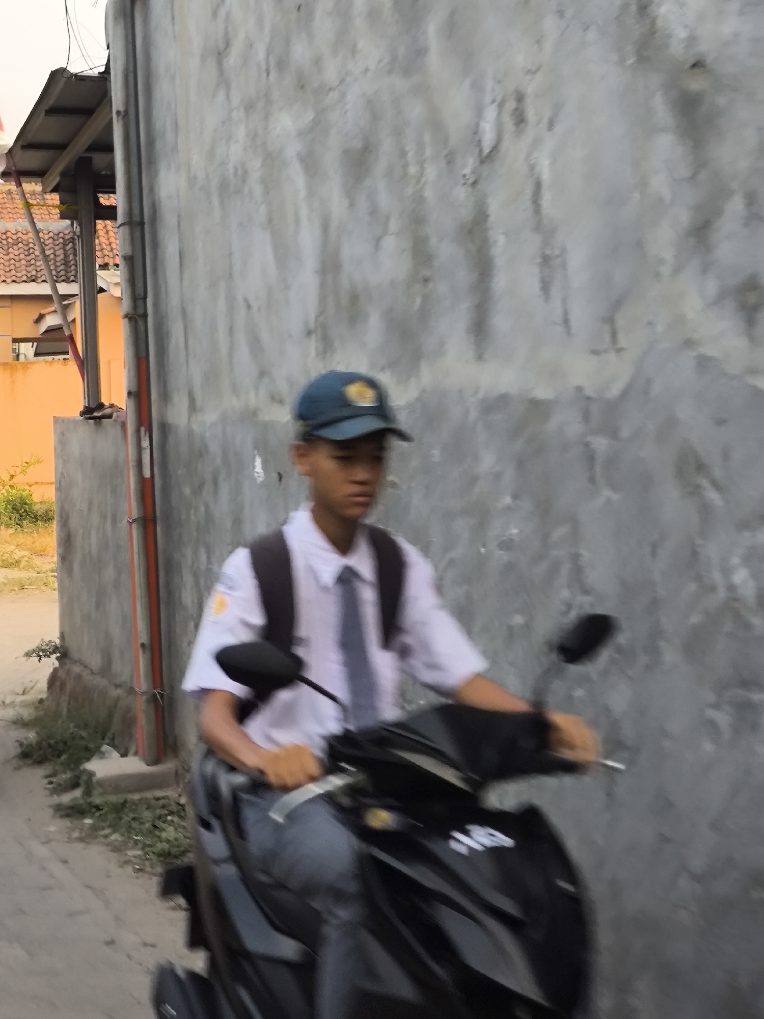 A young male student wearing a white shirt, gray pants, a blue cap, and a backpack rides a black motorcycle down a narrow street in Cilegon, Indonesia. The student is in motion, and the image is slightly blurred due to the movement. The background consists of a rough concrete wall on the right and a building with orange walls and a tiled roof on the left. There is also some vegetation along the side of the street. The lighting suggests it is daytime.