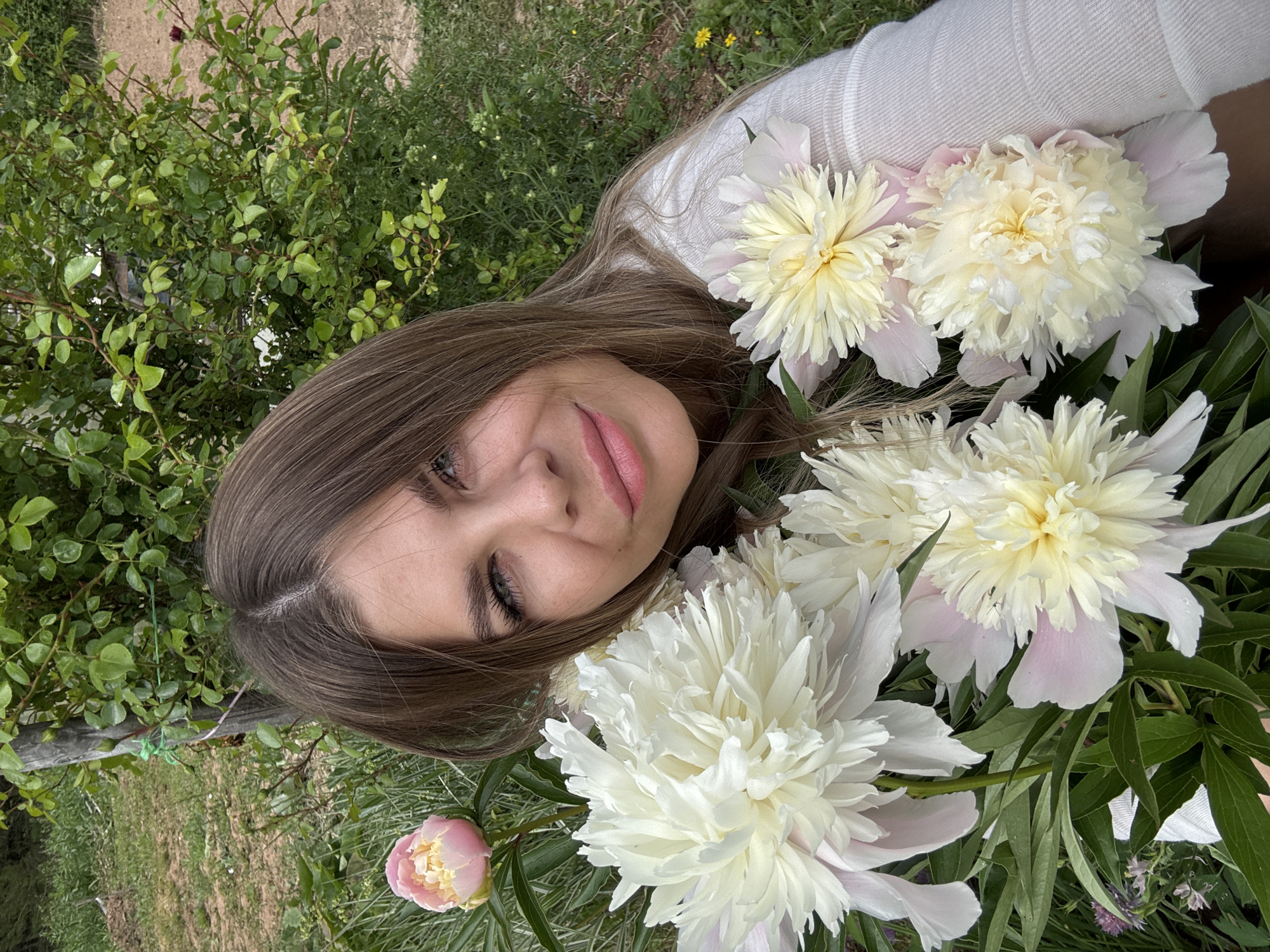 A woman with long, brown hair is taking a selfie outdoors, surrounded by blooming peonies. She is wearing a white, long-sleeved shirt. Several large, white and pink peonies are in the foreground, partially obscuring her face and chest. Behind her, there is lush green foliage and a wooden post. The background appears to be a garden or natural area, with dirt and grass visible. The image does not contain any information indicating the location as Фоки, Russia.