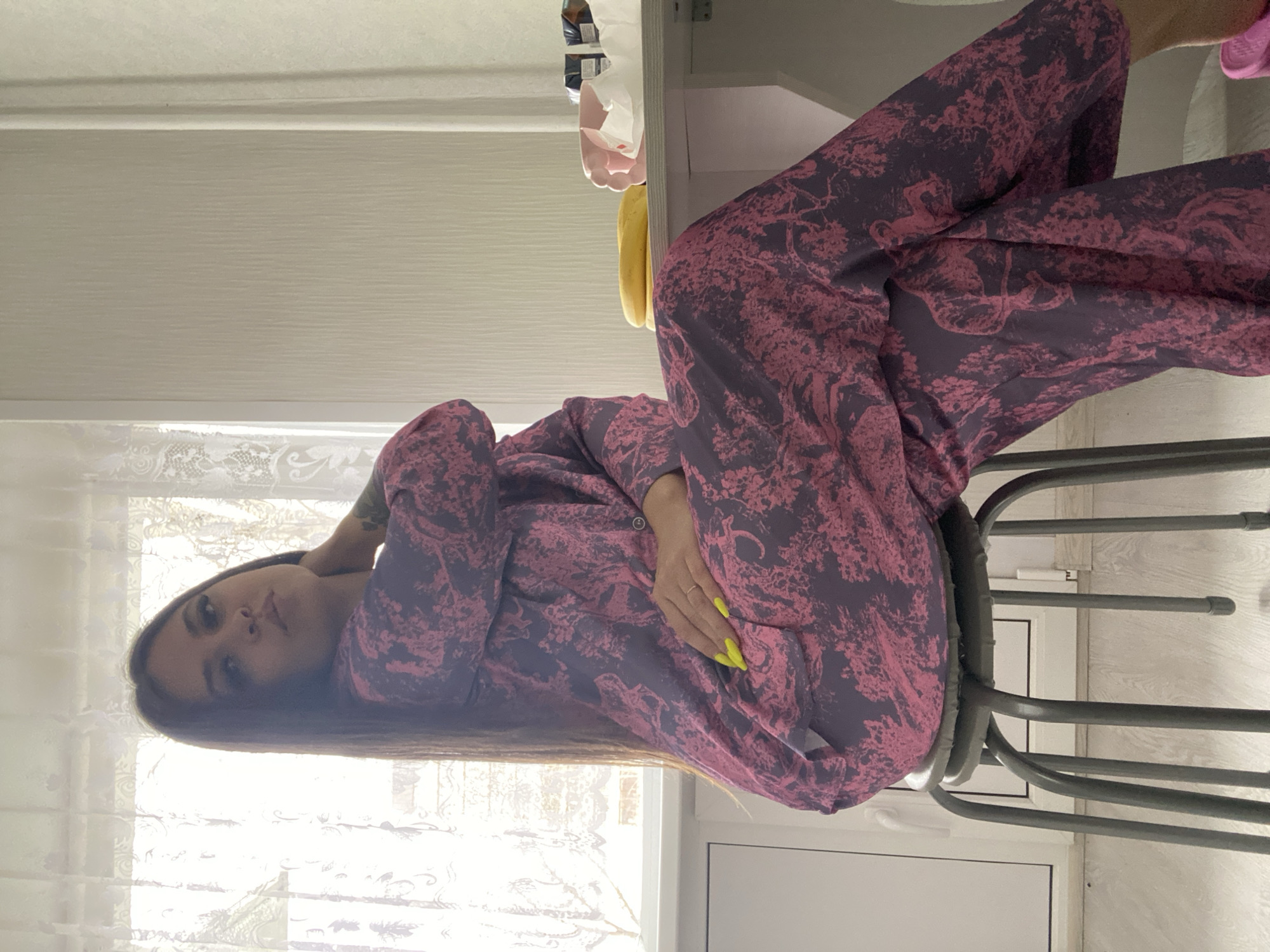 A woman in purple patterned pajamas sits on a metal stool. Her legs are crossed. She is indoors, in a room with a white window and blinds. The specific location is Chaikovsky, Russia.