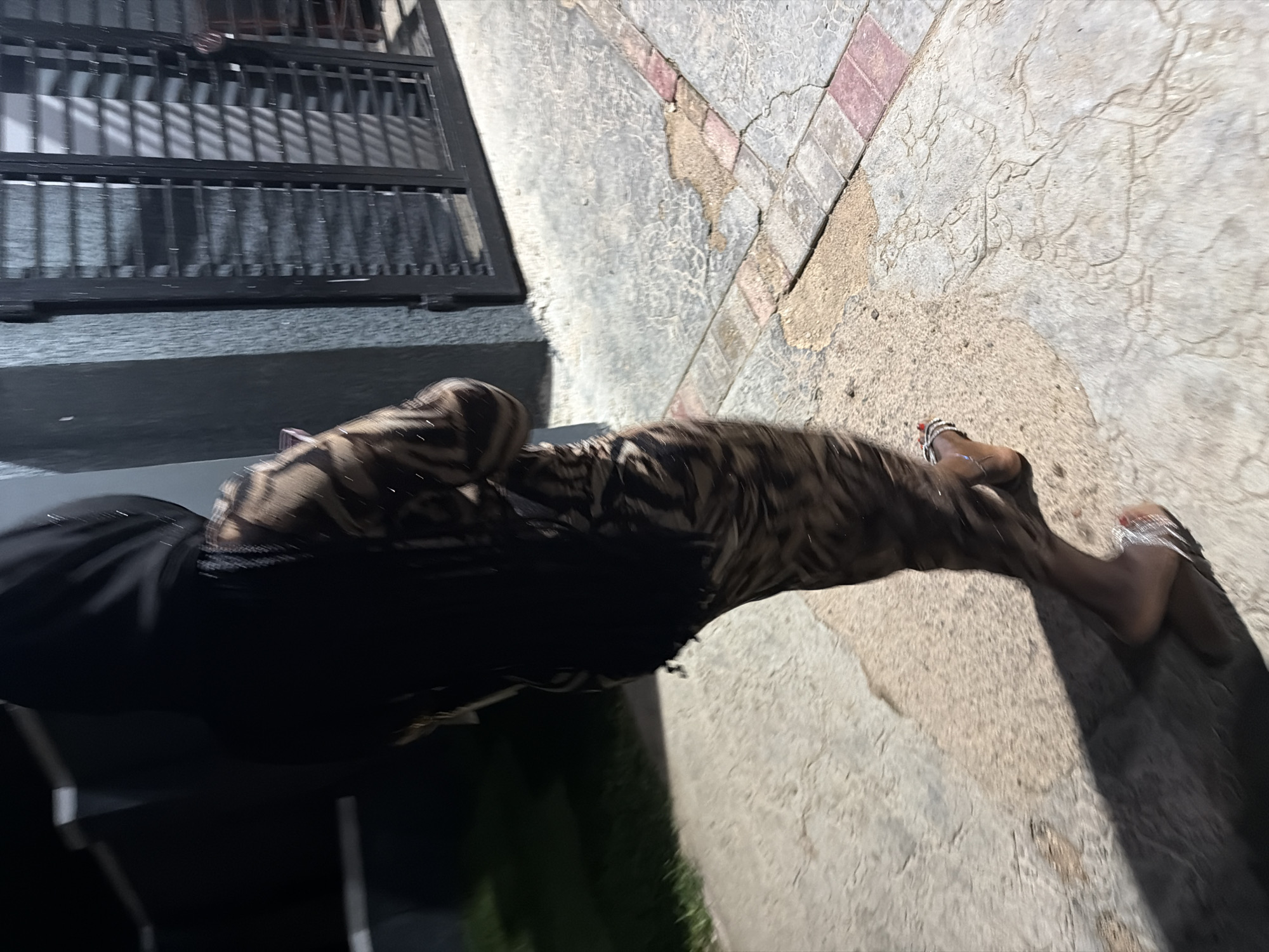 The image shows a person's legs and feet in the foreground, walking on a textured concrete surface. The person is wearing black pants and shoes with a metallic design on the ankles. The pants have a tiger-striped pattern. The scene appears to be outdoors, possibly in a paved area with some grass visible in the lower left corner. A dark metal gate with vertical bars is visible in the background on the left, along with a grey concrete structure. The ground is made of interlocking concrete slabs, some of which are cracked or worn. The lighting suggests it is either early morning or late evening, casting shadows. The location is identified as Bukuru, Nigeria. No text is visible in the image.