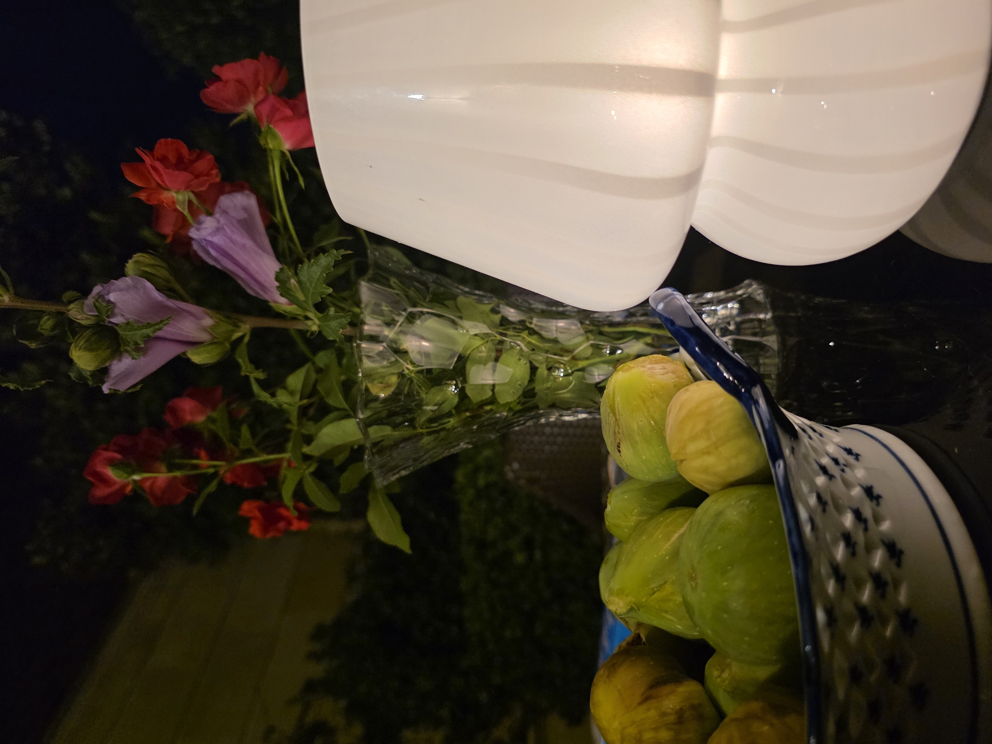 The image depicts an outdoor scene at night, likely in Buzovna, Azerbaijan, with a table set for a casual gathering. In the foreground, a ceramic bowl filled with fresh figs is placed on a dark, reflective surface. The figs are a mix of green and yellowish-brown hues, suggesting ripeness. To the right of the bowl, a white, globe-shaped lamp emits a soft, warm glow, illuminating the surrounding area. Behind the figs, a clear glass vase holds a bouquet of vibrant red and pale purple flowers, with lush green leaves. The background is dimly lit, showing dark foliage and the silhouette of a building. The overall impression is one of tranquil evening ambiance, with natural elements and soft lighting creating a serene atmosphere.