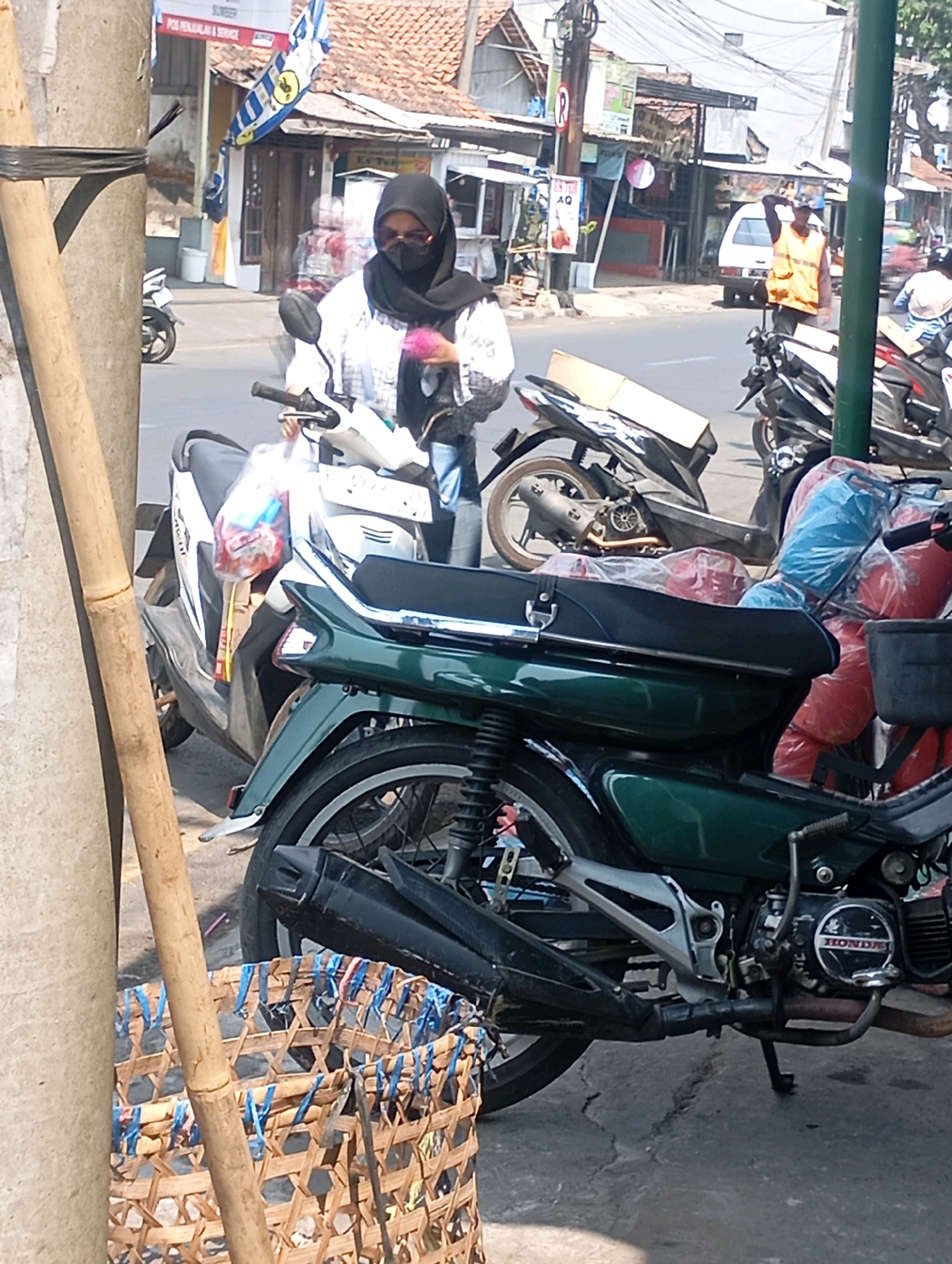 A woman wearing a black hijab and a white patterned shirt is on a white motorcycle. She is wearing sunglasses and a black face mask. She is holding a pink fuzzy object in her right hand. The motorcycle is parked on a street in front of a building. There are other motorcycles parked behind her. In the background, there are buildings, power lines, and a white van. The street is paved and there is a bamboo pole in the foreground. The scene appears to be in a commercial or residential area in Sumber, Indonesia.